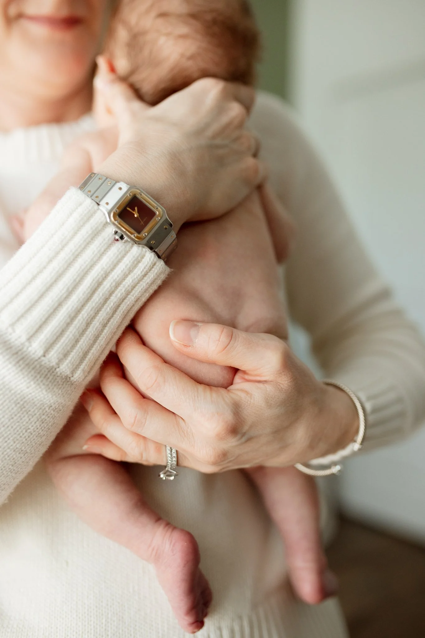 Close-up of a woman holding a newborn baby, with her hand gently supporting the baby's head, wearing a watch and a ring, while the baby is lying on her chest.