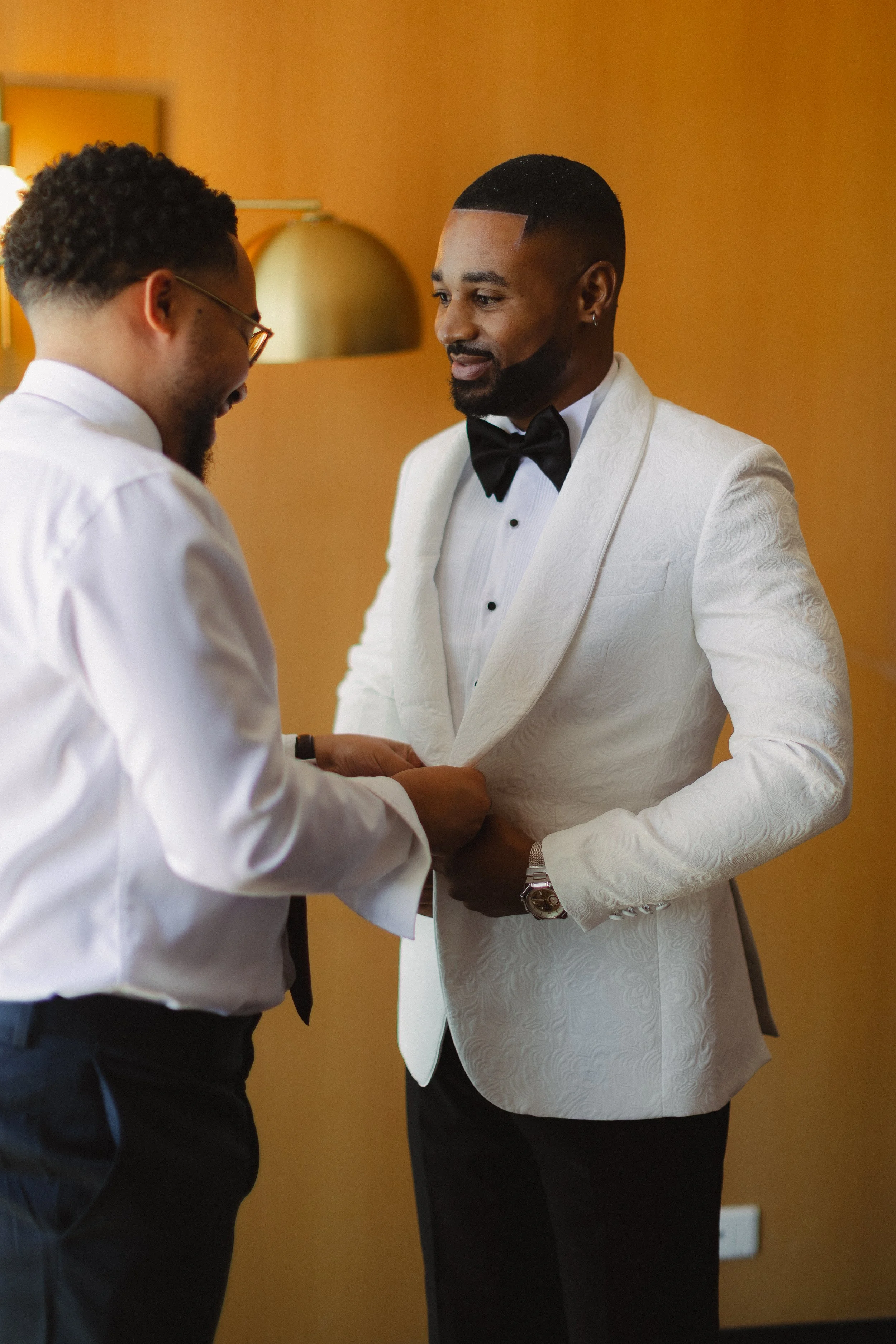 Deux hommes en costume blanc lors d'une cérémonie, échangeant un anneau dans une pièce avec une lumière chaleureuse.