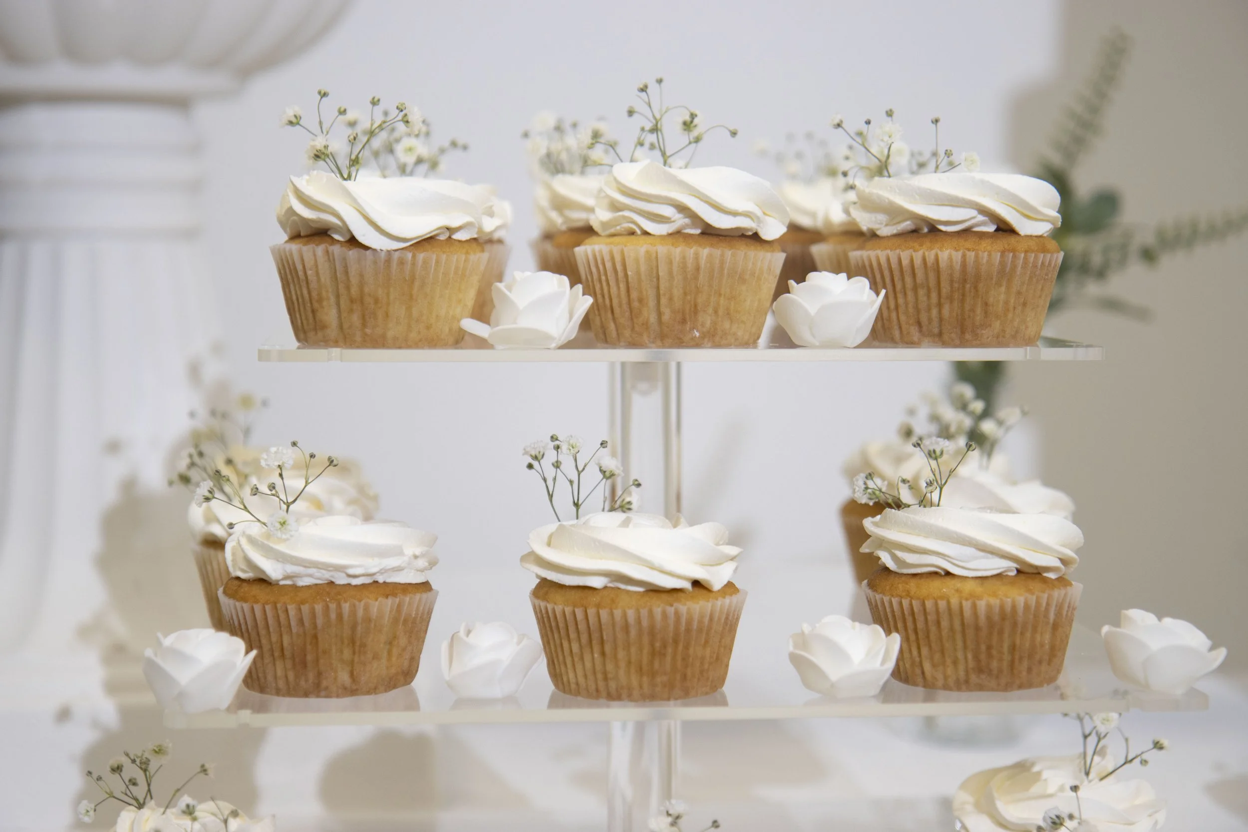 Cupcakes à la vanille avec de la crème blanche décorés de petites fleurs blanches, disposés sur un présentoir à plusieurs niveaux.