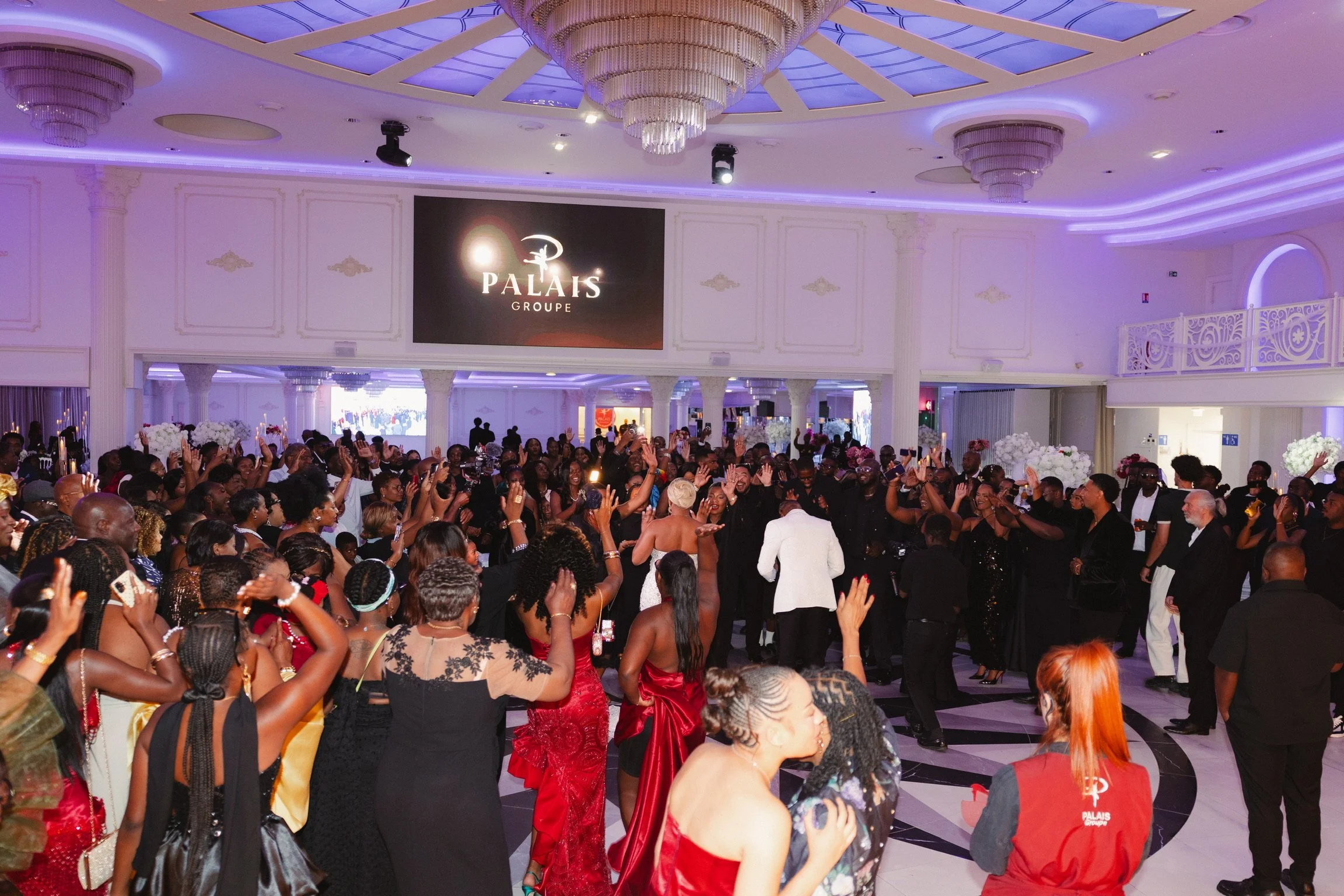 Un grand rassemblement de personnes dans une salle élégante avec un plafond orné de lustres chandeliers, la majorité portent des vêtements de soirée. Il y a un écran avec le logo 'PALAIS GROUPE' au mur, et toutes les personnes semblent s'amuser et participer à l'événement.