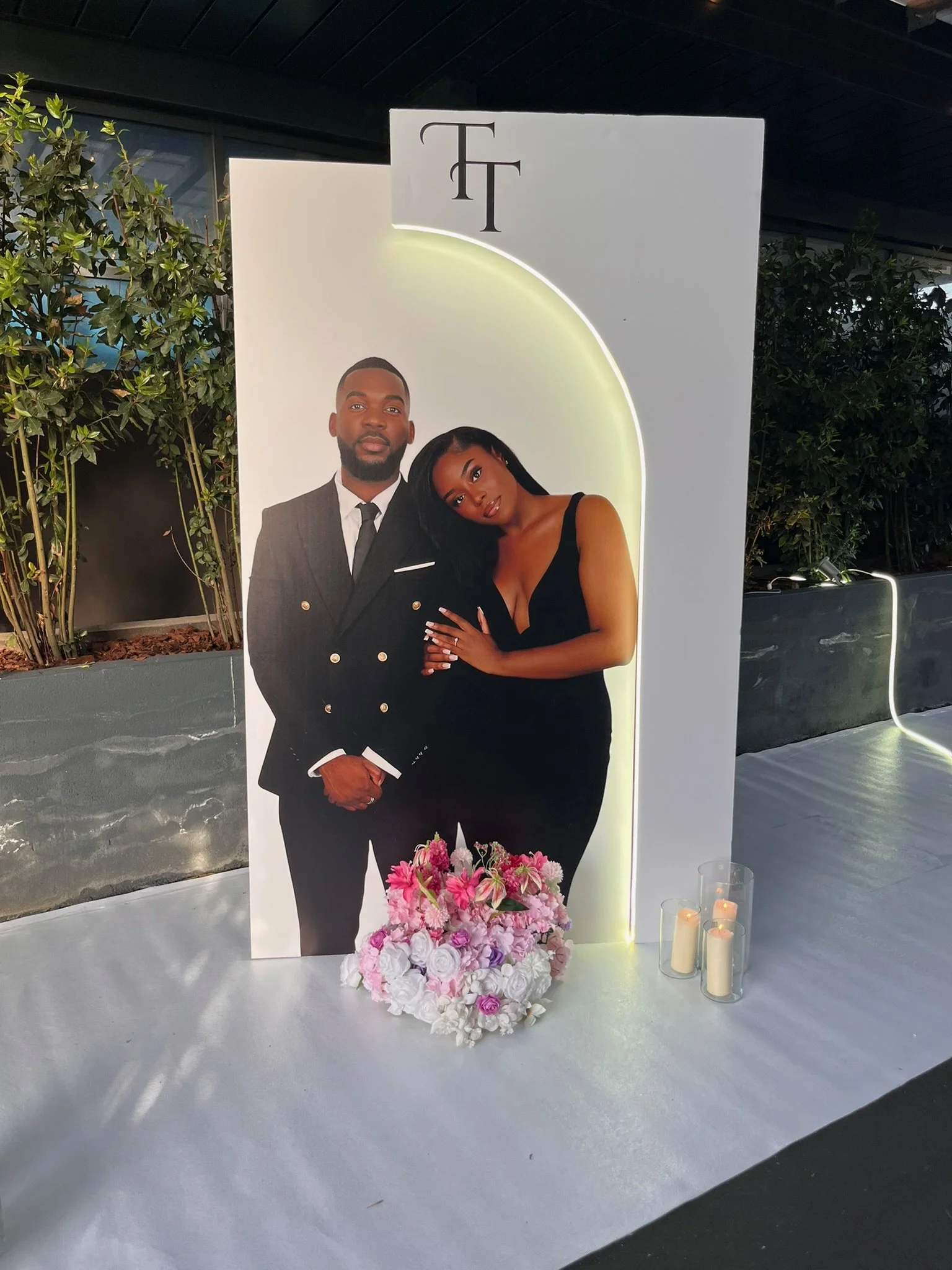 Un panneau décoratif avec une photo de couple lors d'un mariage, orné de fleurs roses et blanches, avec des bougies à côté.