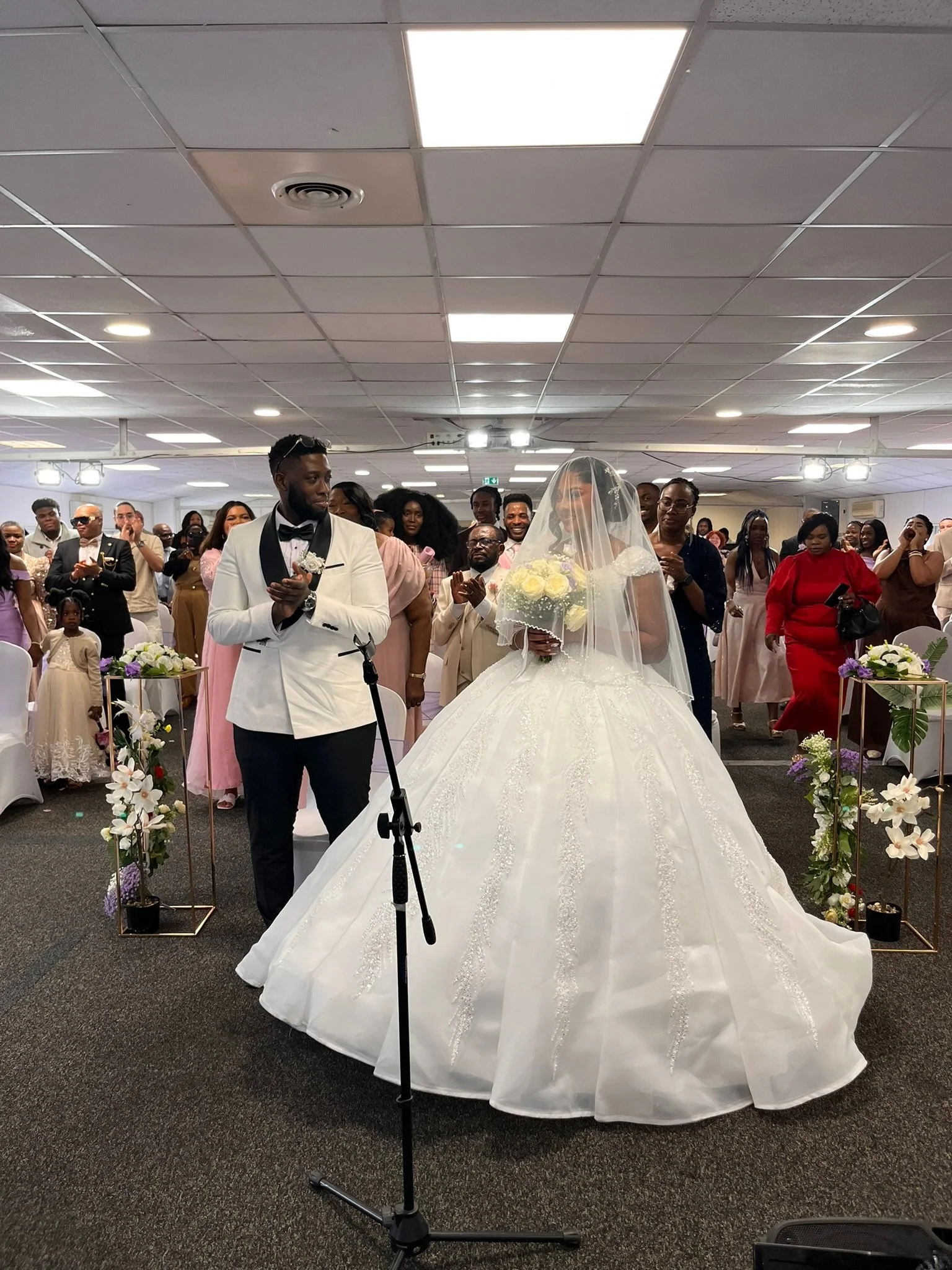 Un couple lors de leur mariage, le peu visible à cause du voile de la mariée, entouré de leurs invités dans une salle de réception. La mariée porte une robe de mariée blanche et un voile, tenant un bouquet de fleurs. Le marié porte un costume blanc avec des boutons noirs et un nœud papillon noir.