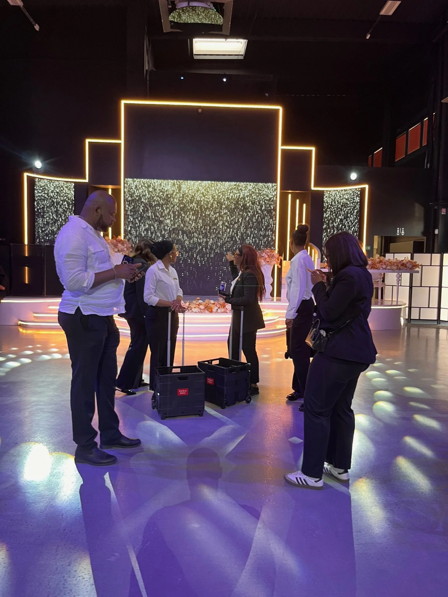 Groupe de personnes dans un espace intérieur, apparemment lors d'une cérémonie ou d'un événement, avec un décor lumineux en arrière-plan et des personnes qui semblent attendre ou participer à une activité.