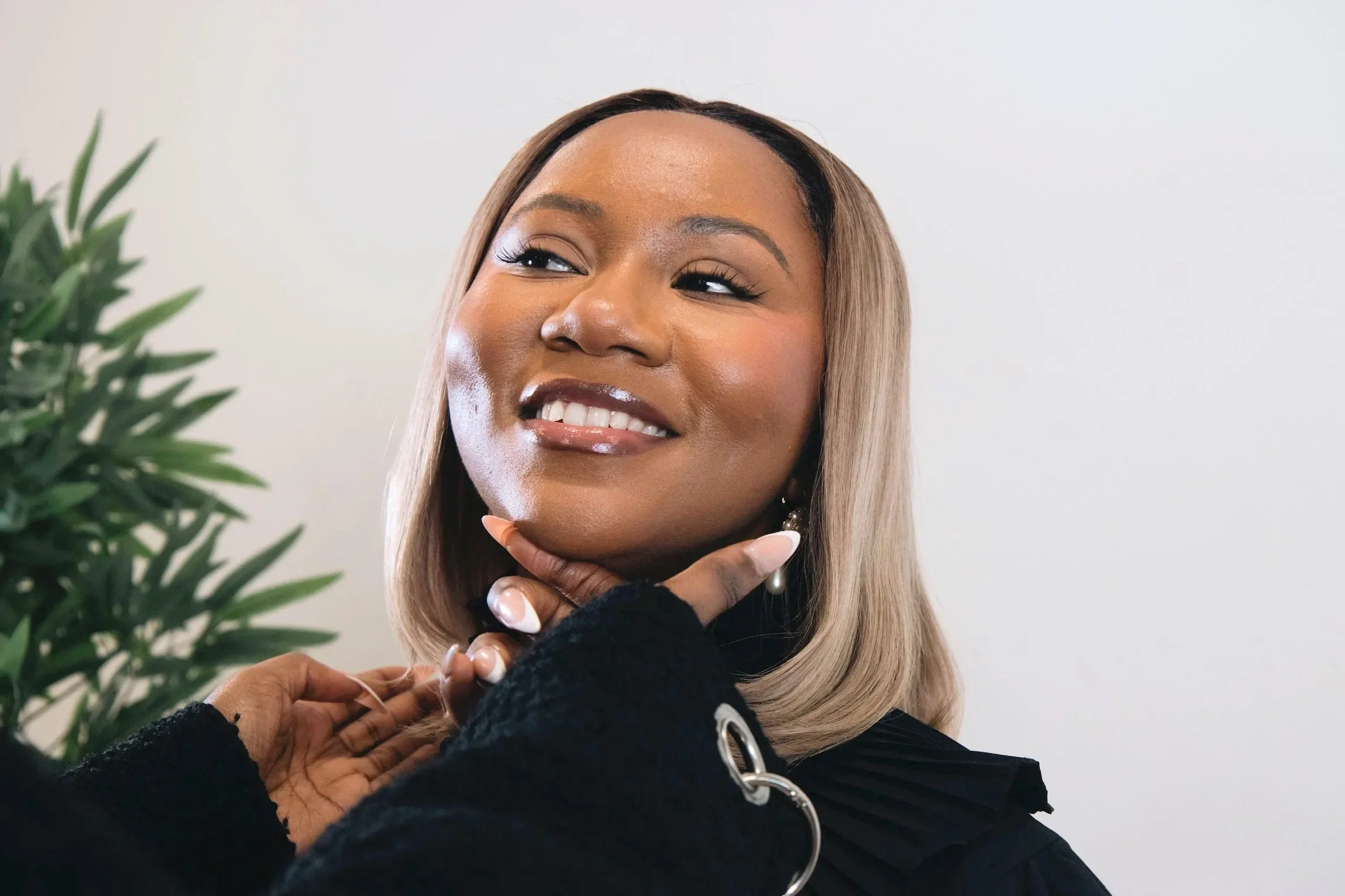 Une femme souriante avec des cheveux mi-longs blonds et un trait de maquillage léger, portant une veste noire et des boucles d'oreilles pendantes, touche son cou avec une main, se trouvant devant un mur blanc et une plante verte à gauche.
