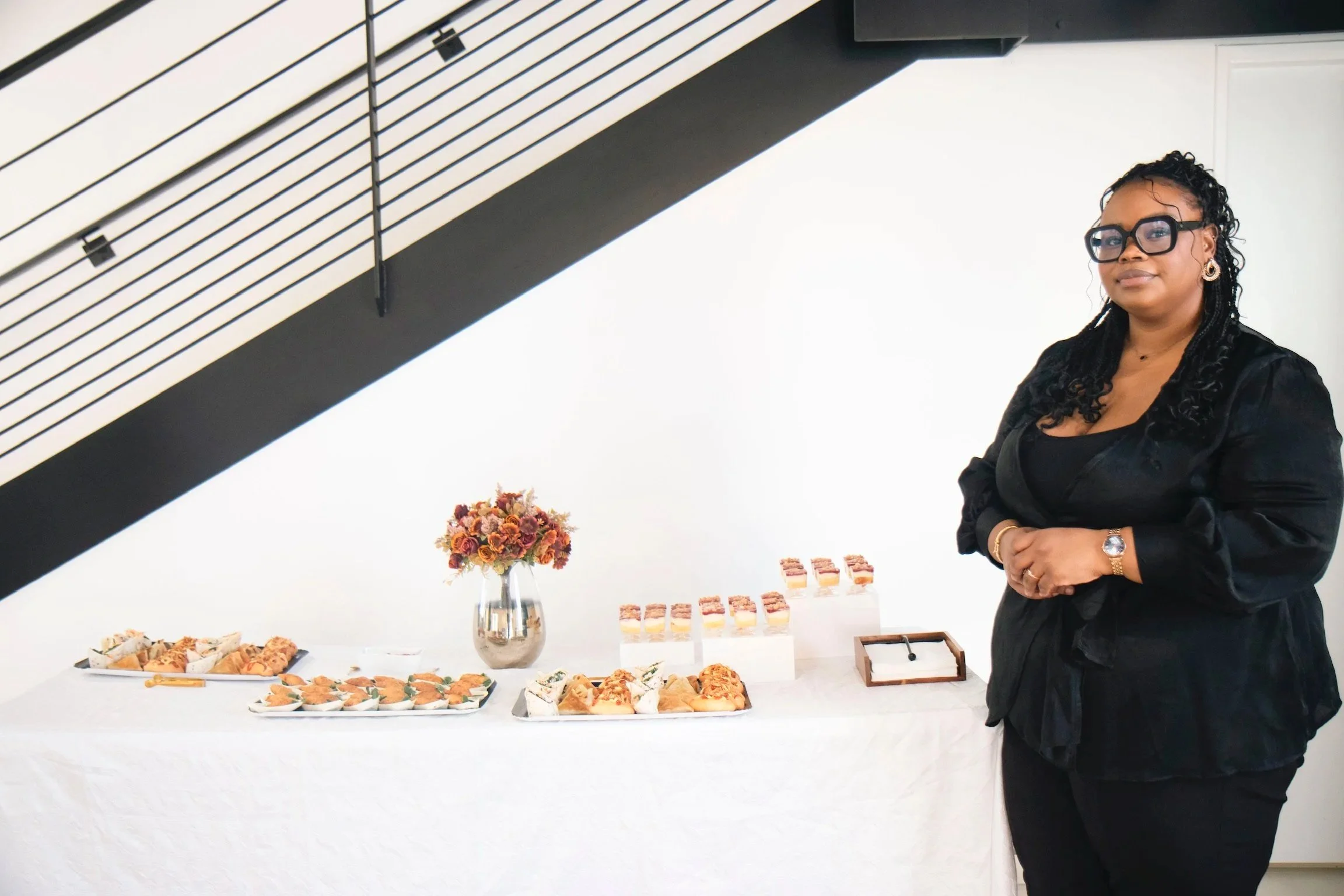 Une femme habillée en noir se tient à côté d'une table avec des amuse-gueules et un vase de fleurs, dans une salle moderne.