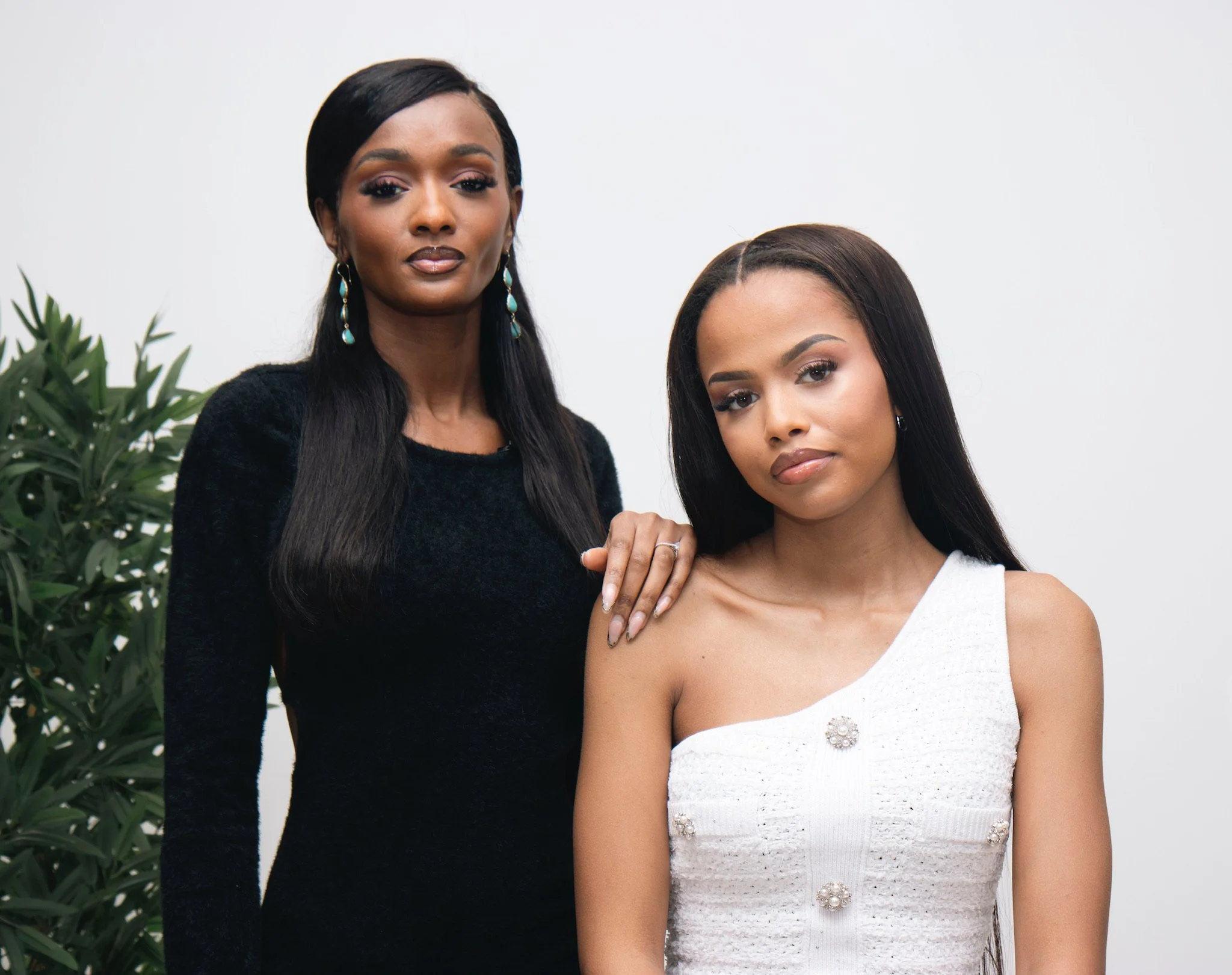 Deux femmes posant pour une photo, une avec une main sur l'épaule de l'autre, dans un décor intérieur avec une plante verte en arrière-plan.