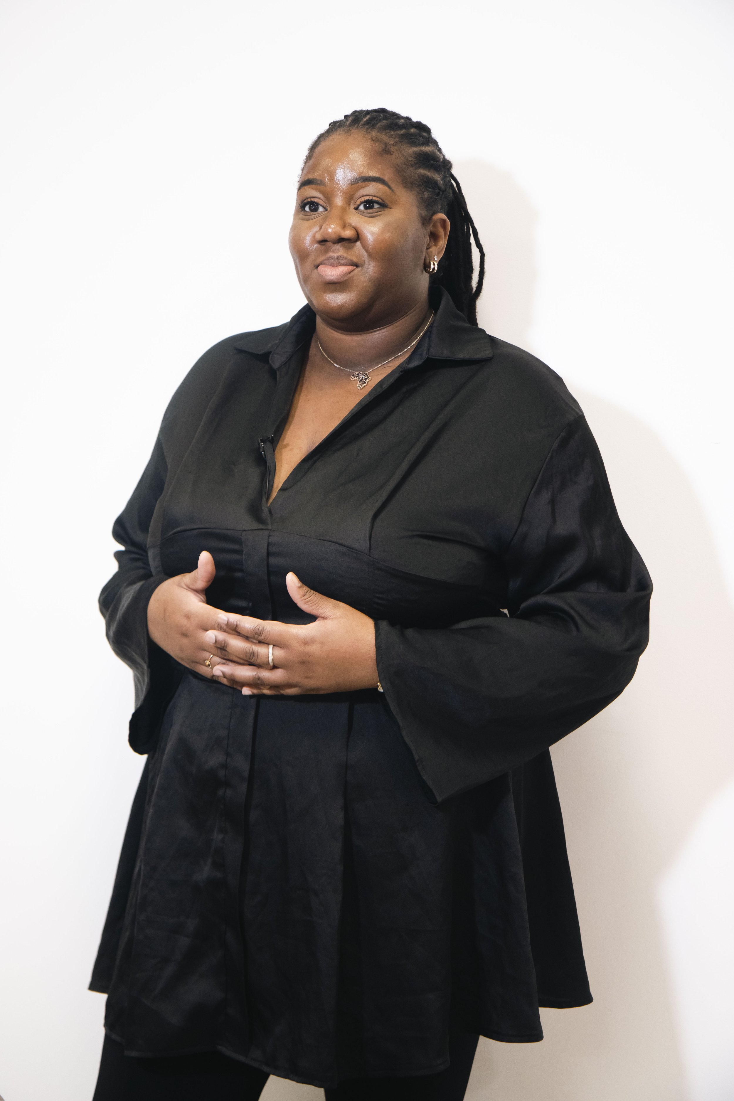 Femme noire portant une robe noire, se tenant contre un mur blanc, regardant légèrement à gauche, avec un sourire léger, ignore le contexte de l'image.