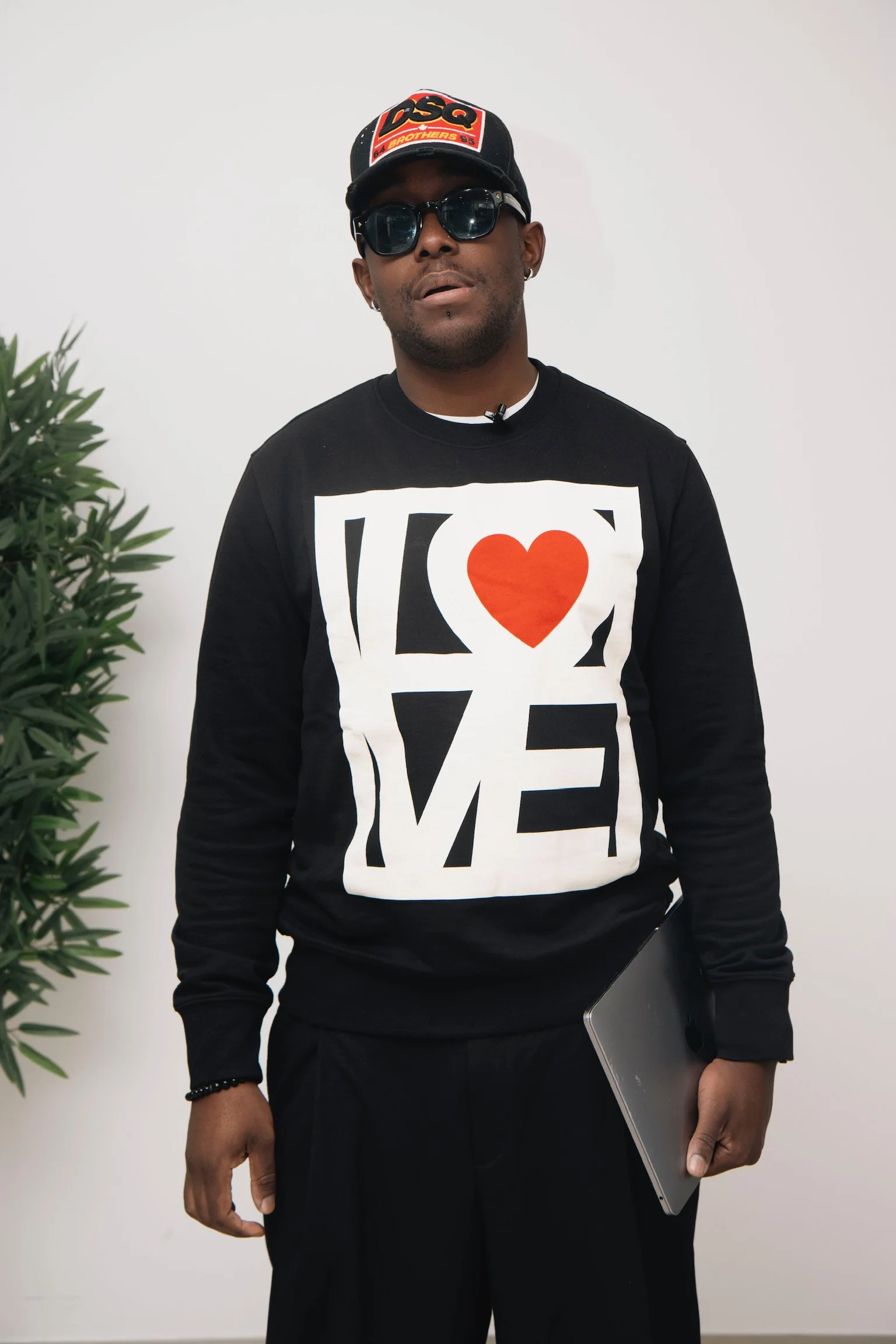 Un homme portant un sweat noir avec l'inscription LOVE et un cœur rouge, des lunettes de soleil, un chapeau avec le logo LSQ, et tenant un ordinateur portable, debout près d'une plante verte, contre un mur blanc.