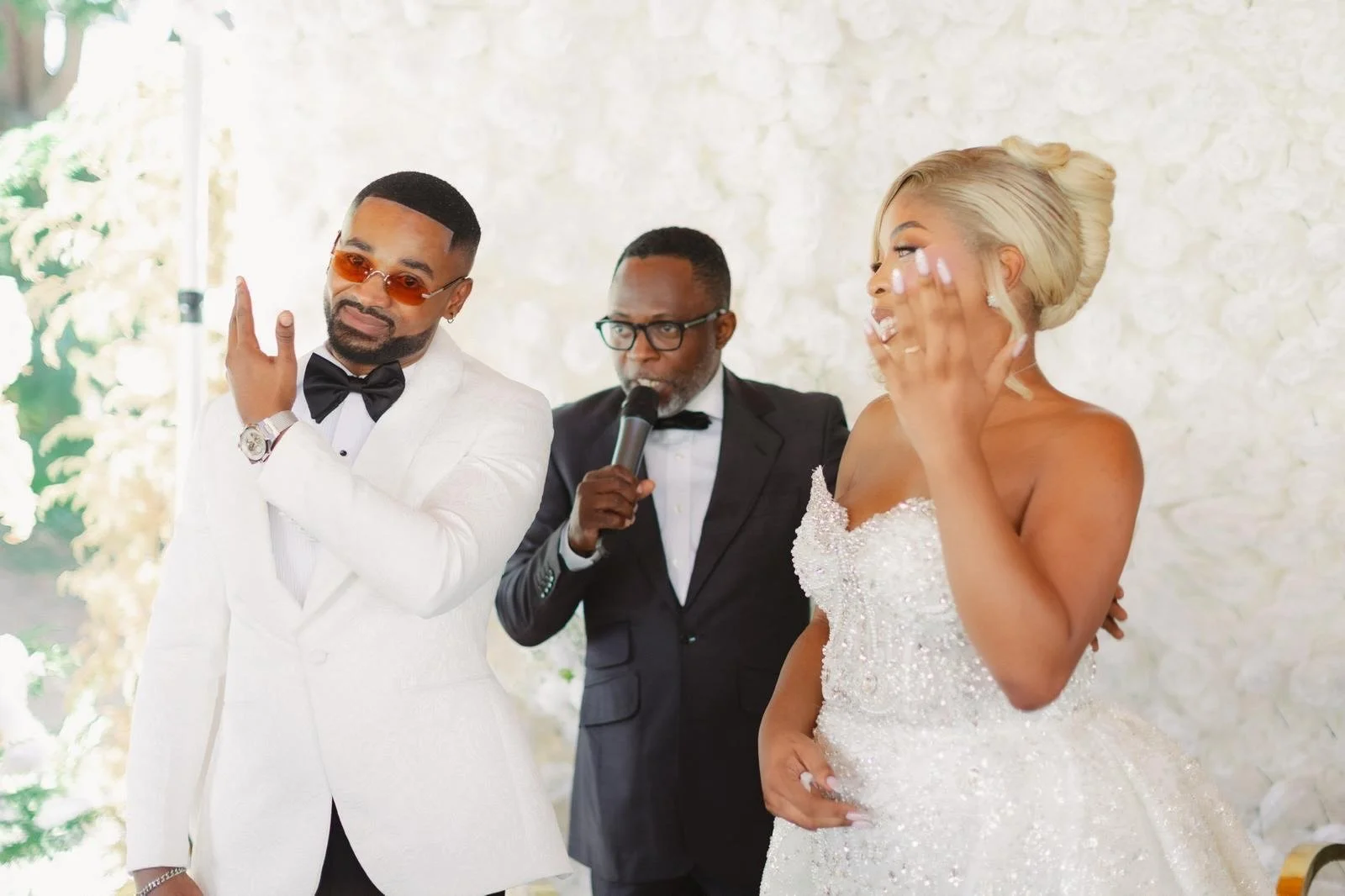 Un couple lors de leur mariage, avec un officiant en costume noir, une femme en robe de mariée blanche et un homme en costume blanc avec des lunettes de soleil orange.