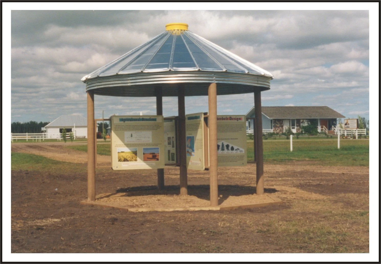 An outdoor informational kiosk with a metal roof and wooden support posts, displaying large informational panels about agriculture and design, situated in a field with houses and a white fence in the background under a cloudy sky.