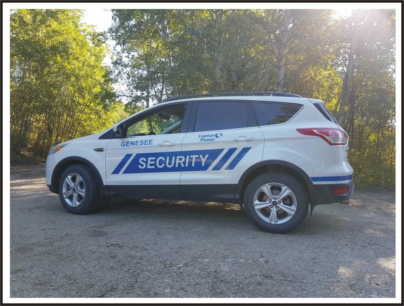 A white security vehicle with blue markings and the words 'Security' and 'Genesee' on the side, parked on a dirt surface surrounded by green trees and sunlight filtering through the branches.