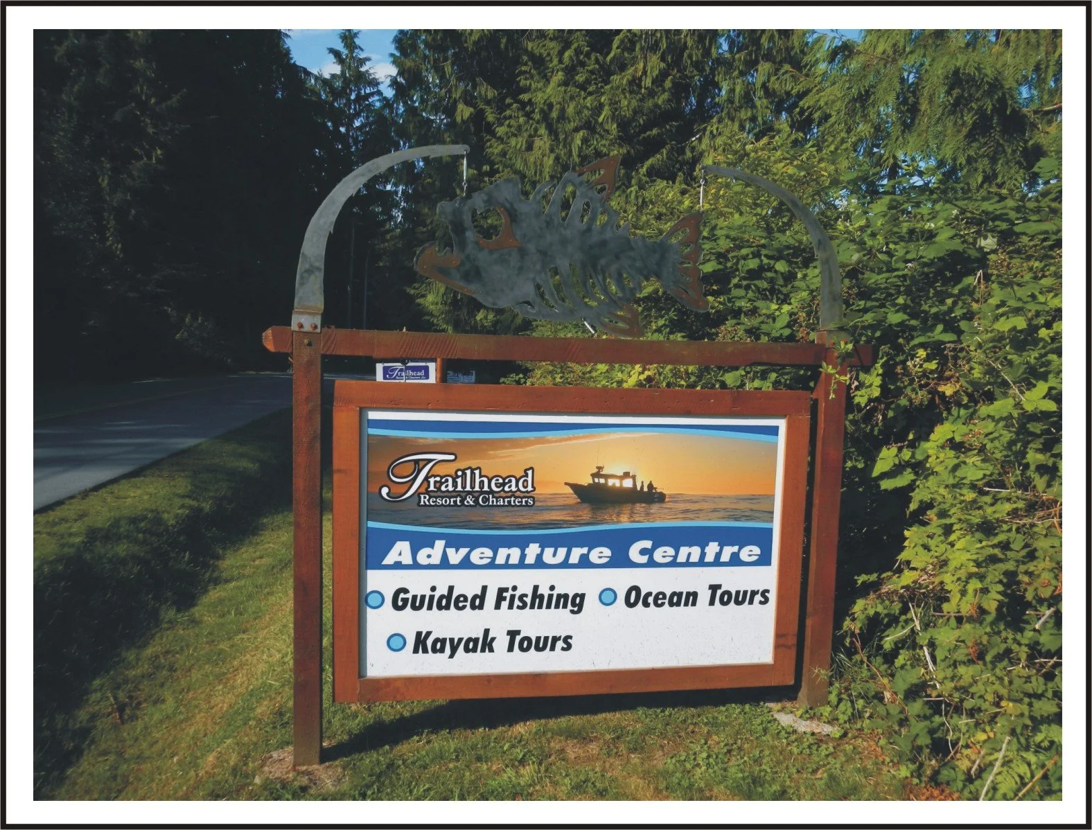 Sign for Trailhead Resort & Charters advertising guided fishing, ocean tours, and kayak tours, with a background of trees and a boat at sunset.