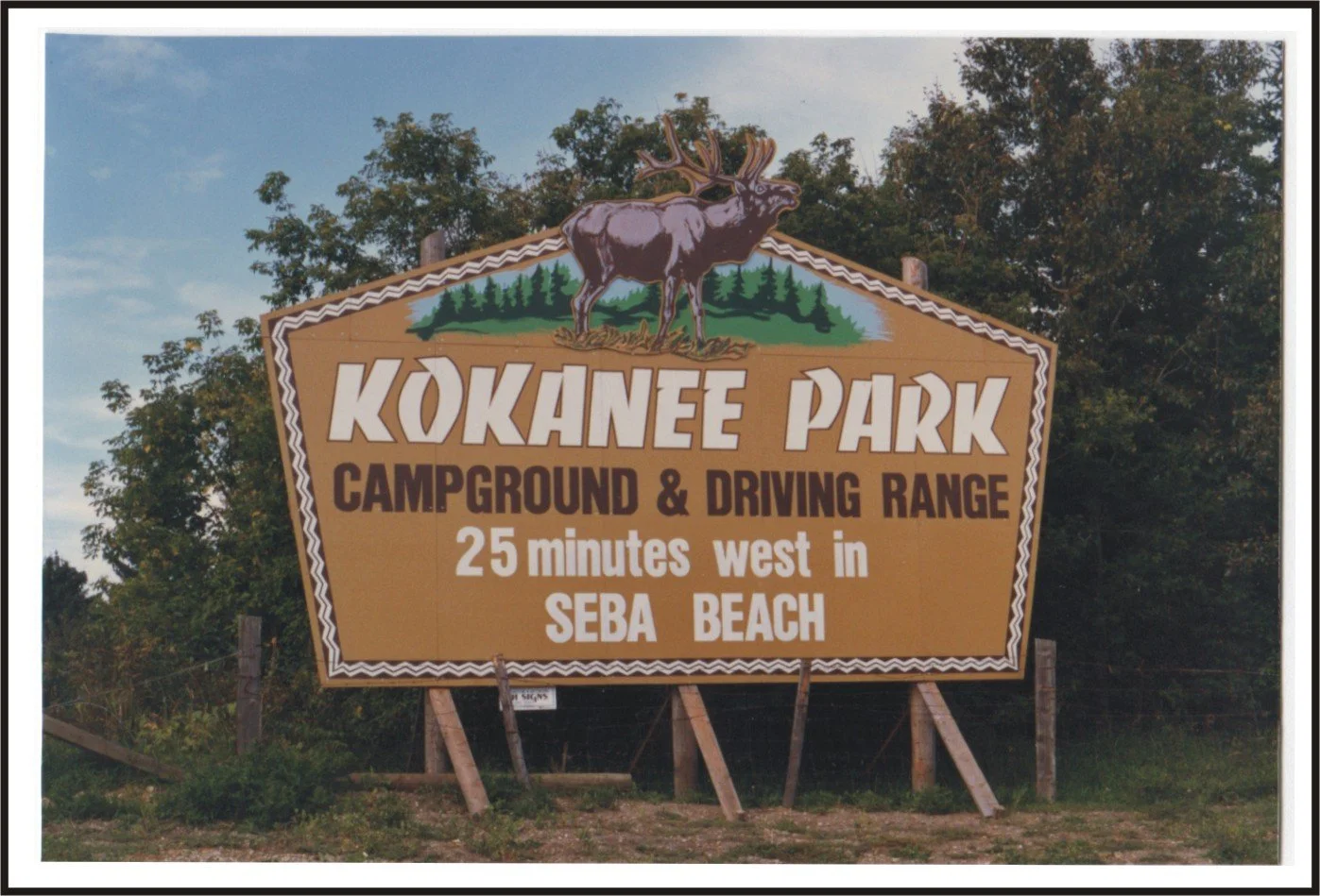 Sign for Kokanee Park indicating campground and driving range, located 25 minutes west in Seba Beach, with trees in background.