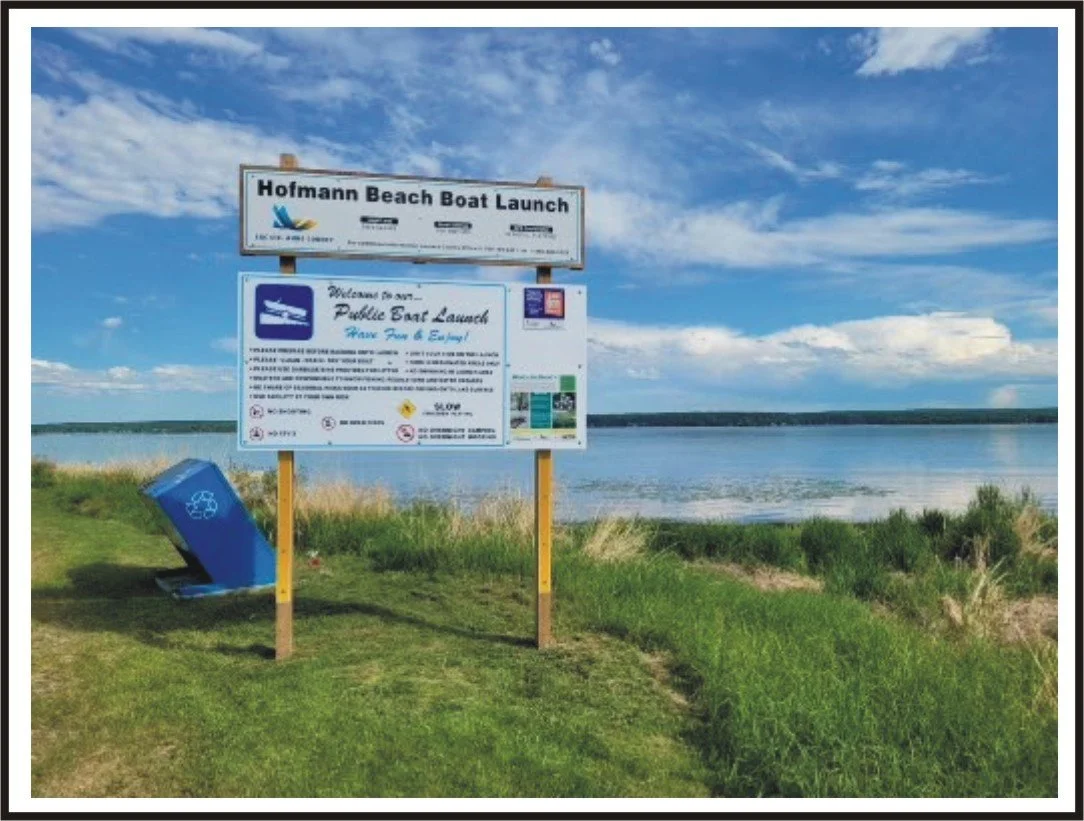 Sign at Hofmann Beach indicating boat launch rules and instructions, with a recycling bin nearby, overlooking a body of water under a blue sky with clouds.