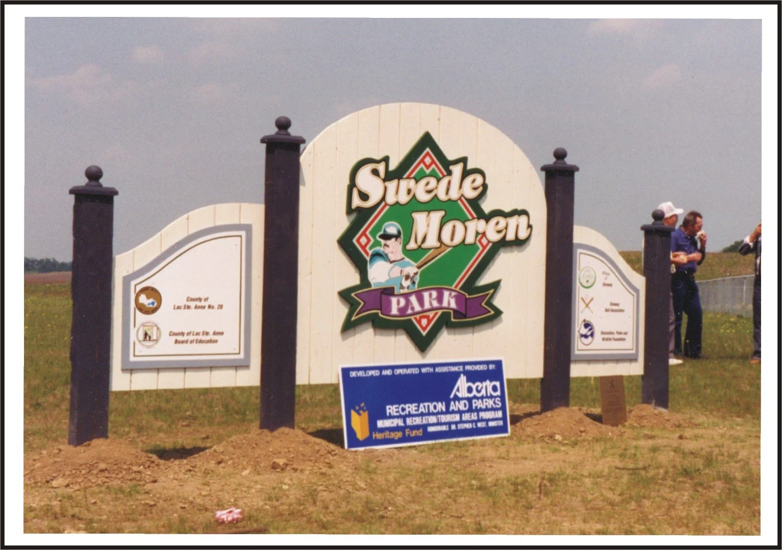Sign for Suede Moran Park with the logo and additional informational signs, and a group of people standing to the right outdoors.