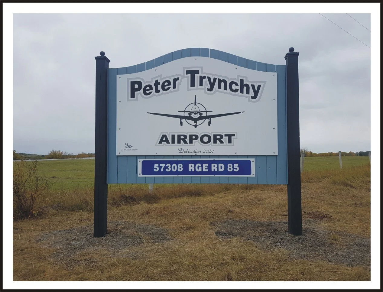 Sign for Peter Trynchy Airport with an airplane graphic, located at 57308 RGE RD 85, in a rural area with grass and overcast sky.