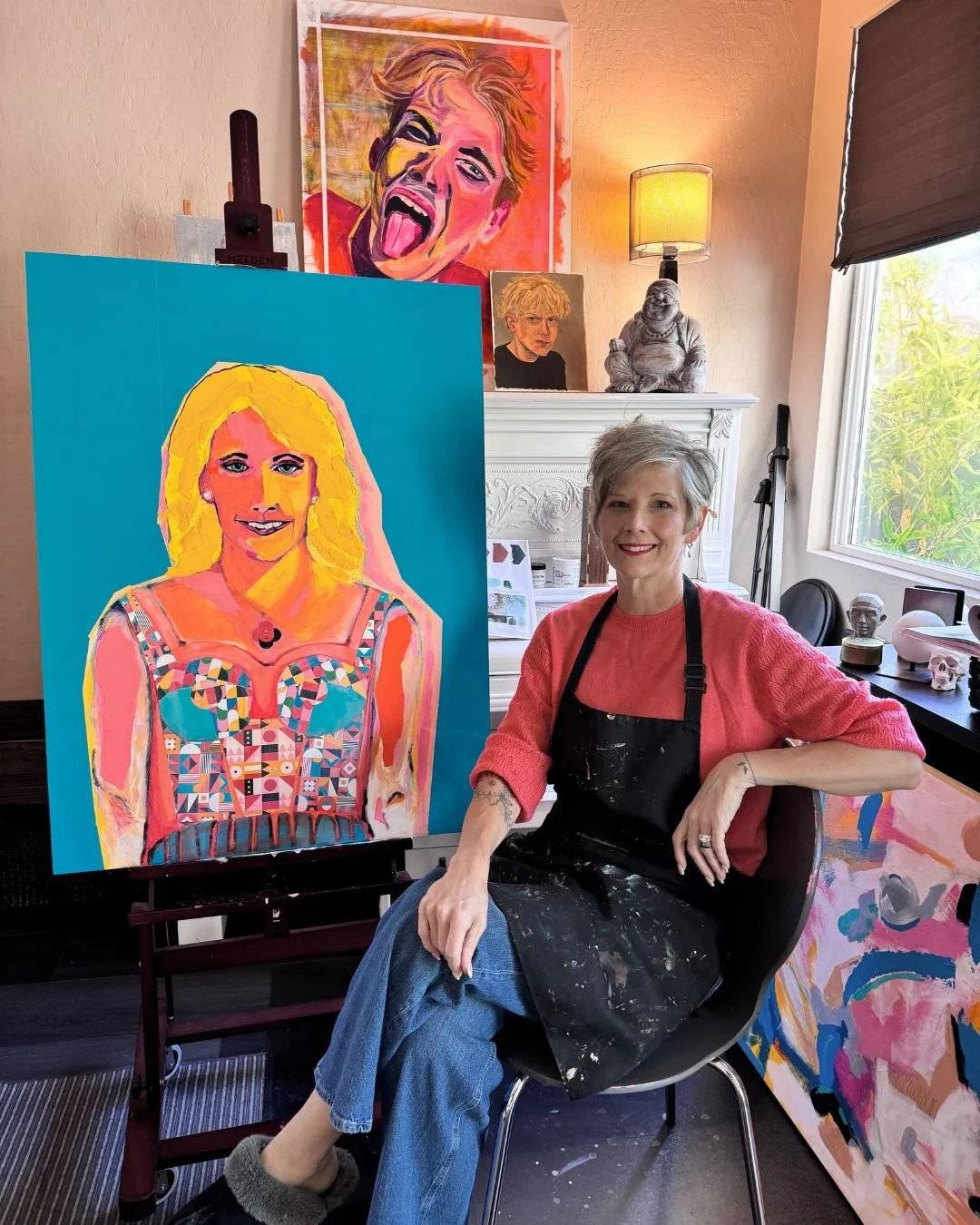 Artist Gina Payne seated in her studio beside a colorful commissioned portrait on an easel