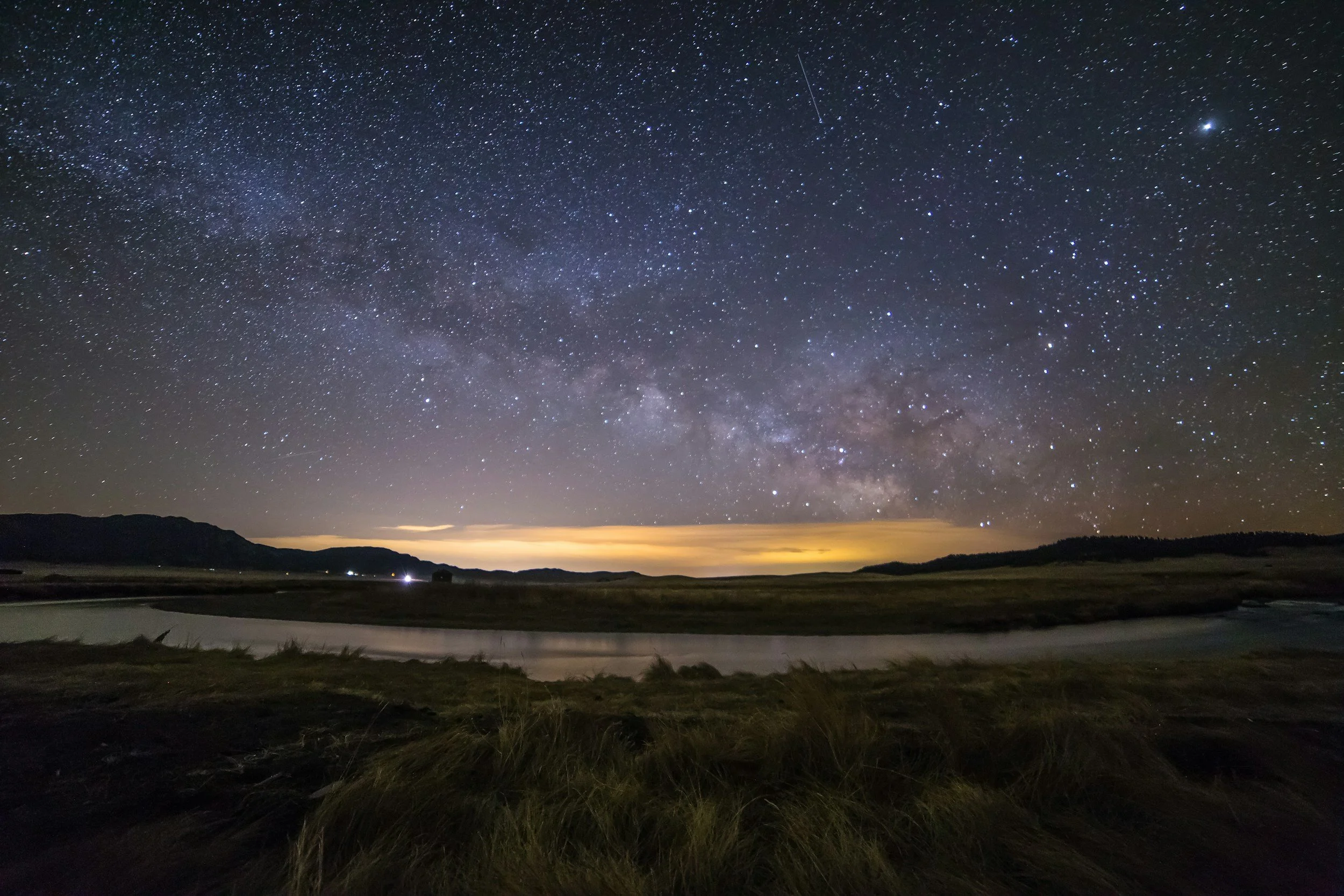 Nighttime landscape with a star-filled sky and the Milky Way galaxy visible, over a calm river and grassy land with distant hills and a faint glow on the horizon.