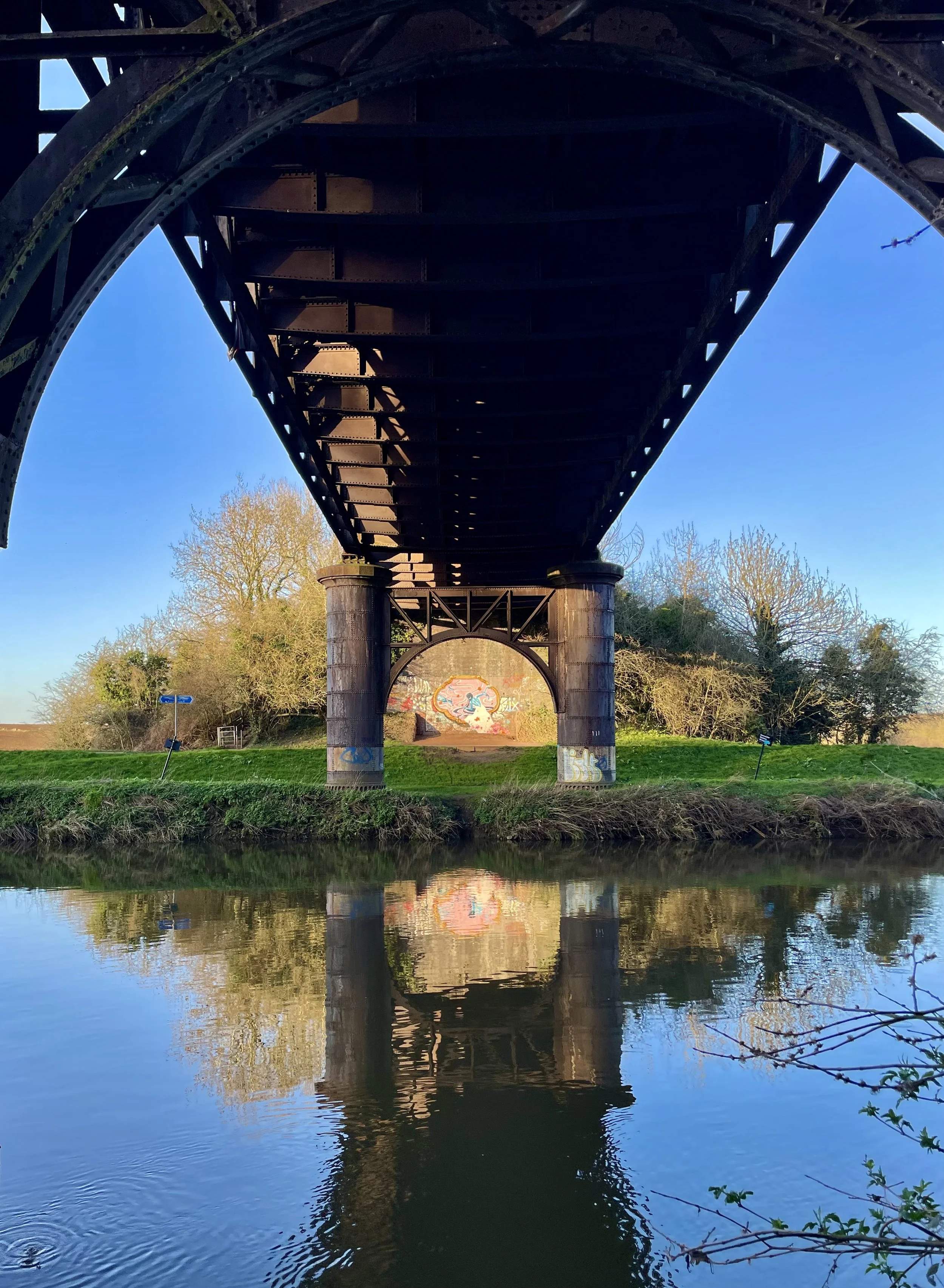 Underneath a bridge over a river, with graffiti on the wall at the back and trees with bare branches around, and clear blue sky reflection on the water.