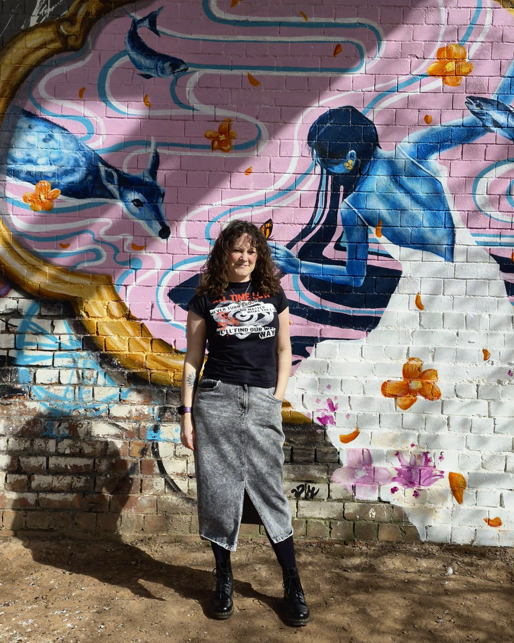 A woman stands in front of a pink brick wall painted with a mural featuring a blue woman with long hair, butterflies, flowers, and swirling patterns.
