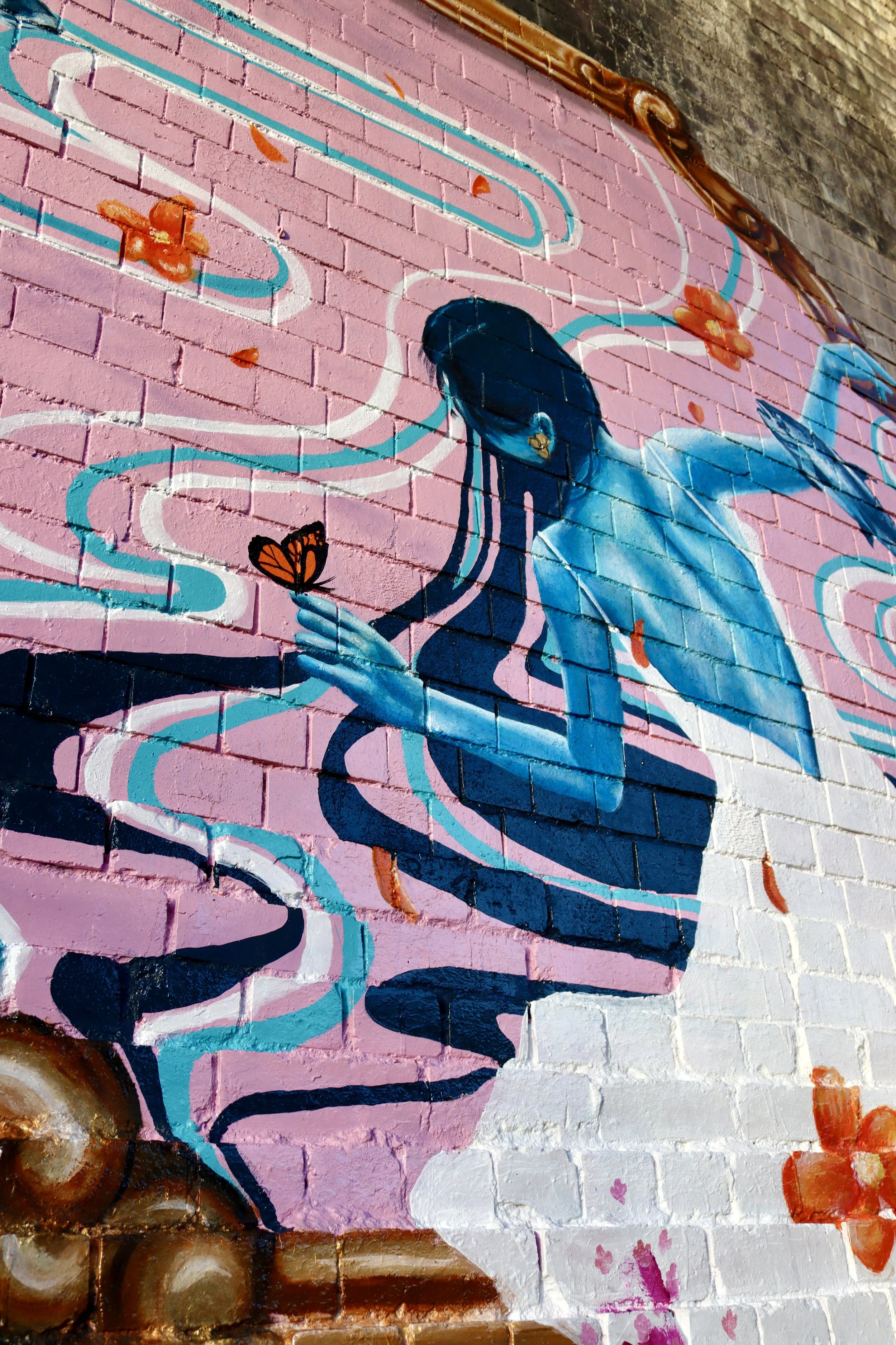 A mural of a woman with blue skin and dark hair, holding a butterfly in her hand, surrounded by pink and orange flowers and flowing lines on a brick wall.