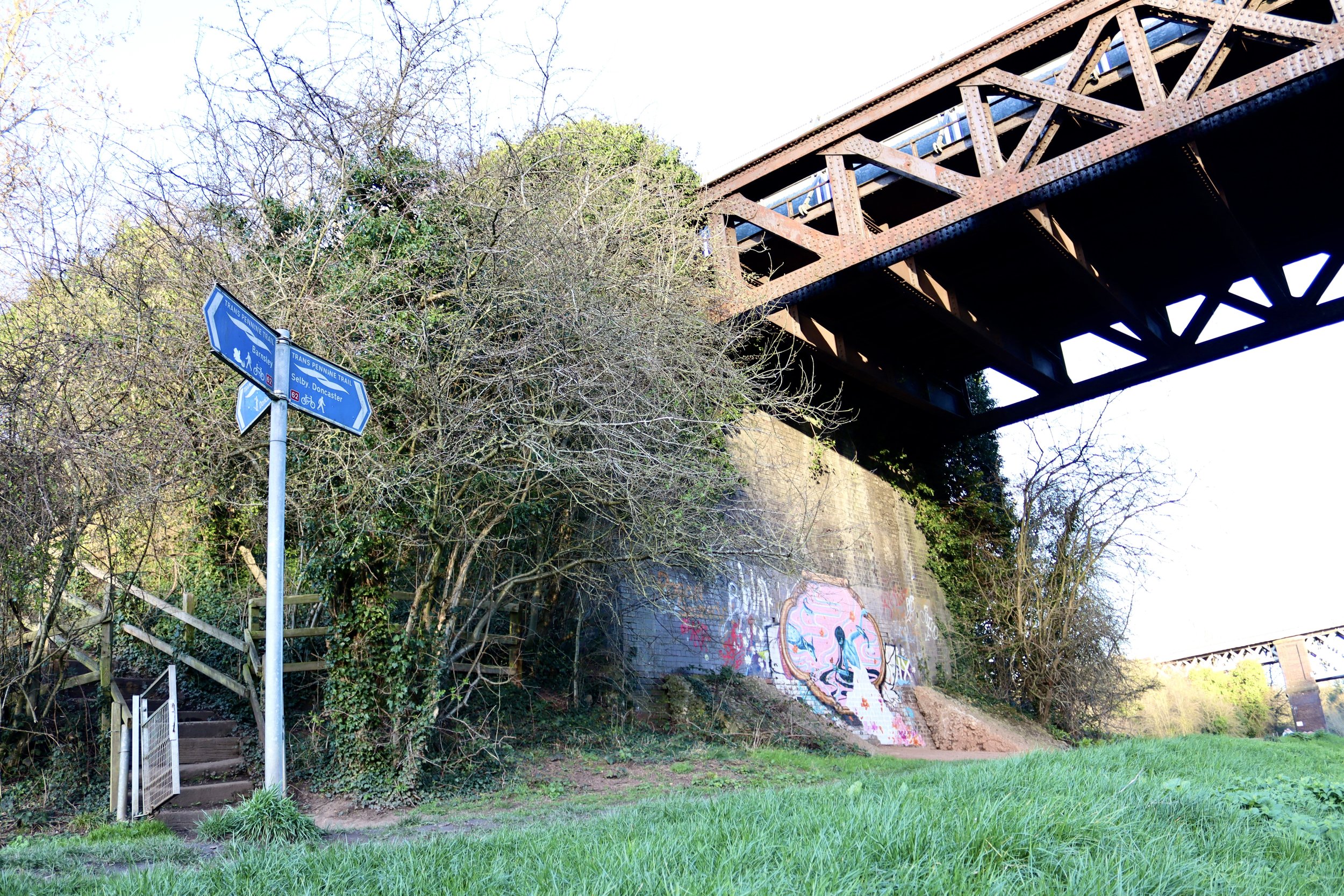 A pathway beneath a railway bridge in Warmsworth, Doncaster, featuring UNFURL's debut mural 'Guardian' on the embankment, trees and a blue way finder sign. 