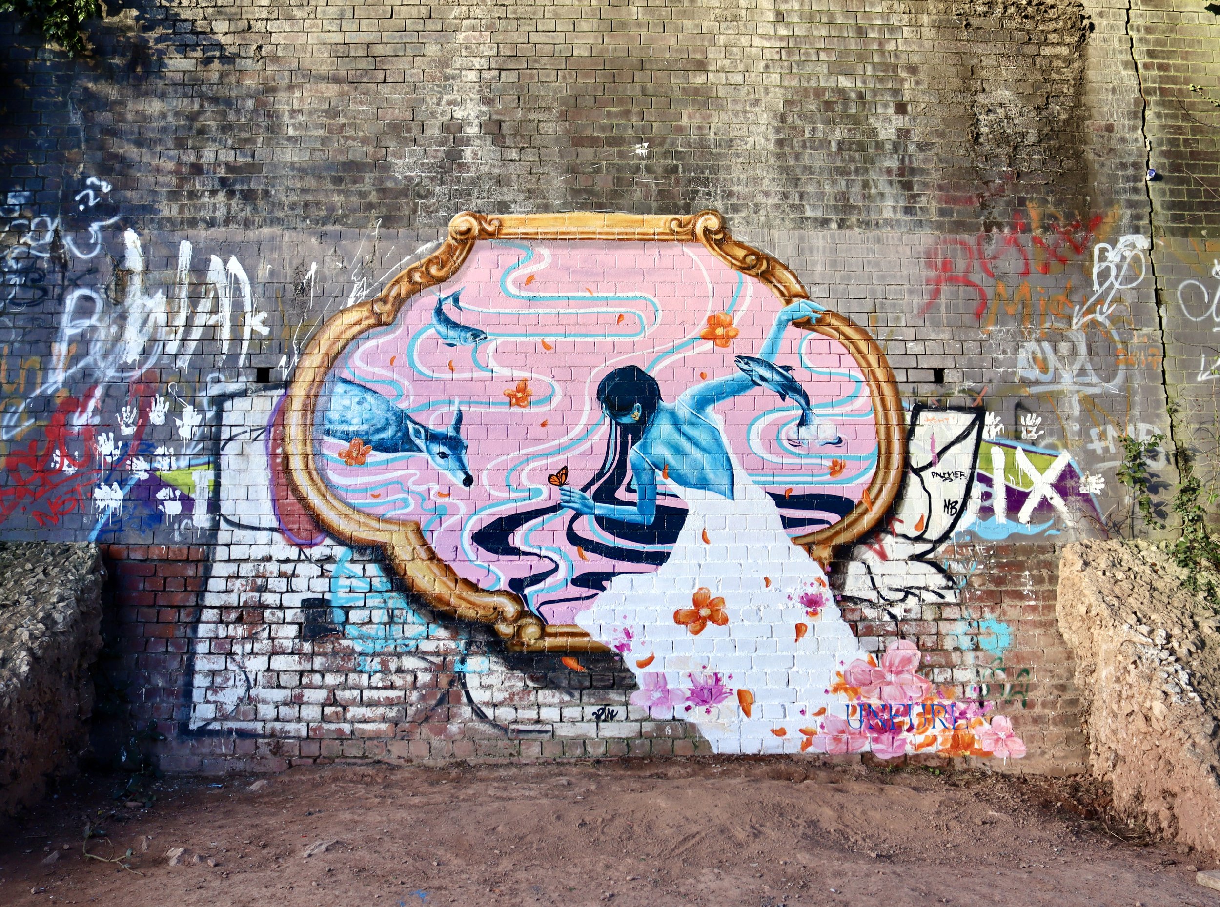 UNFURL's debut mural 'Guardian' Featuring a blue female water spirit within a decorative gold frame painted on a brick wall. The background has pink and blue swirls, orange butterflies, and floral elements.