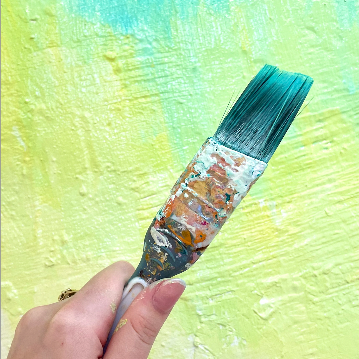 A hand holding a paintbrush covered in multicolored paint, painting a yellow textured wall with a light green tint.