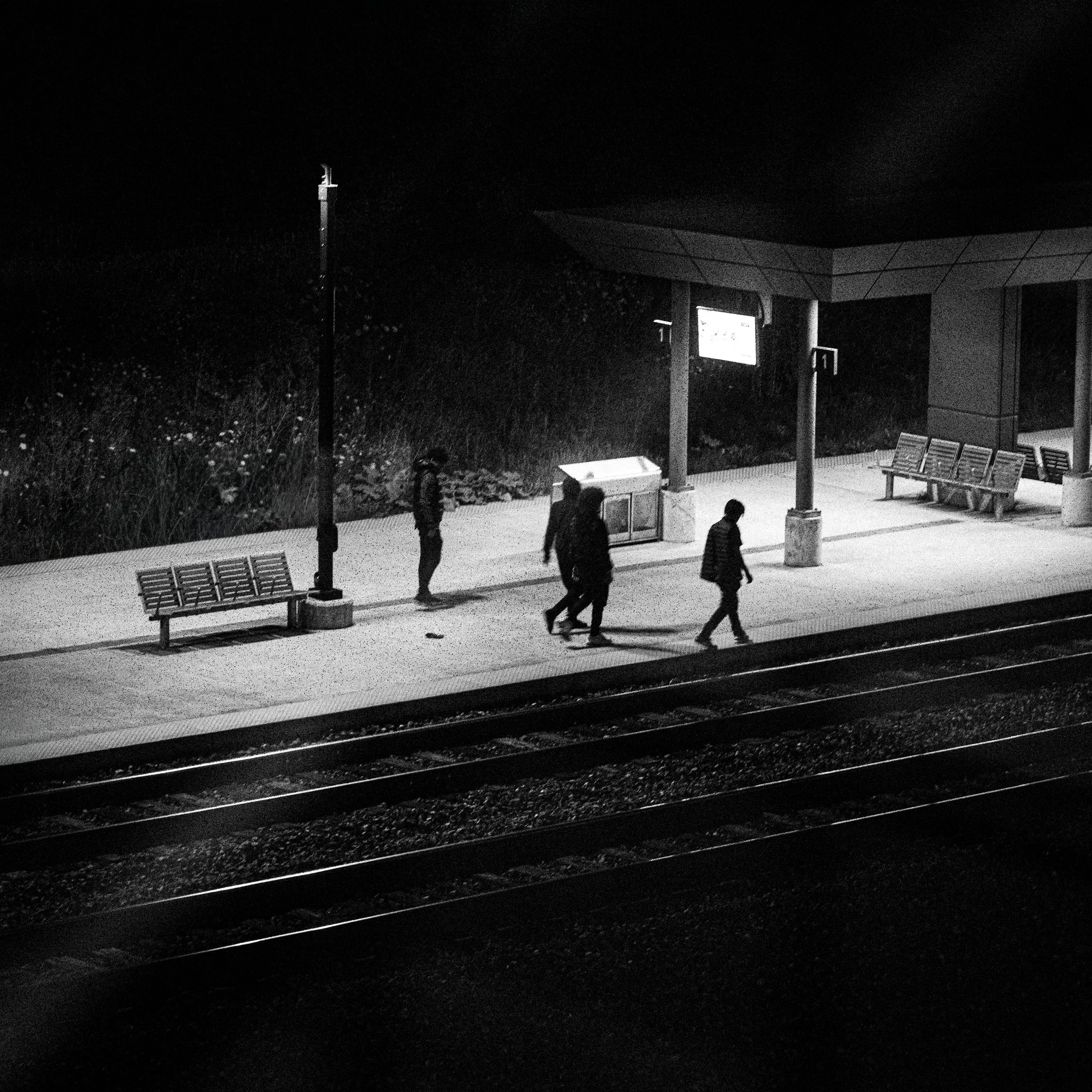Black and white photograph of four people walking on a train station platform at night, with benches, a trash bin, and a shelter. The scene is lit by a single street lamp.