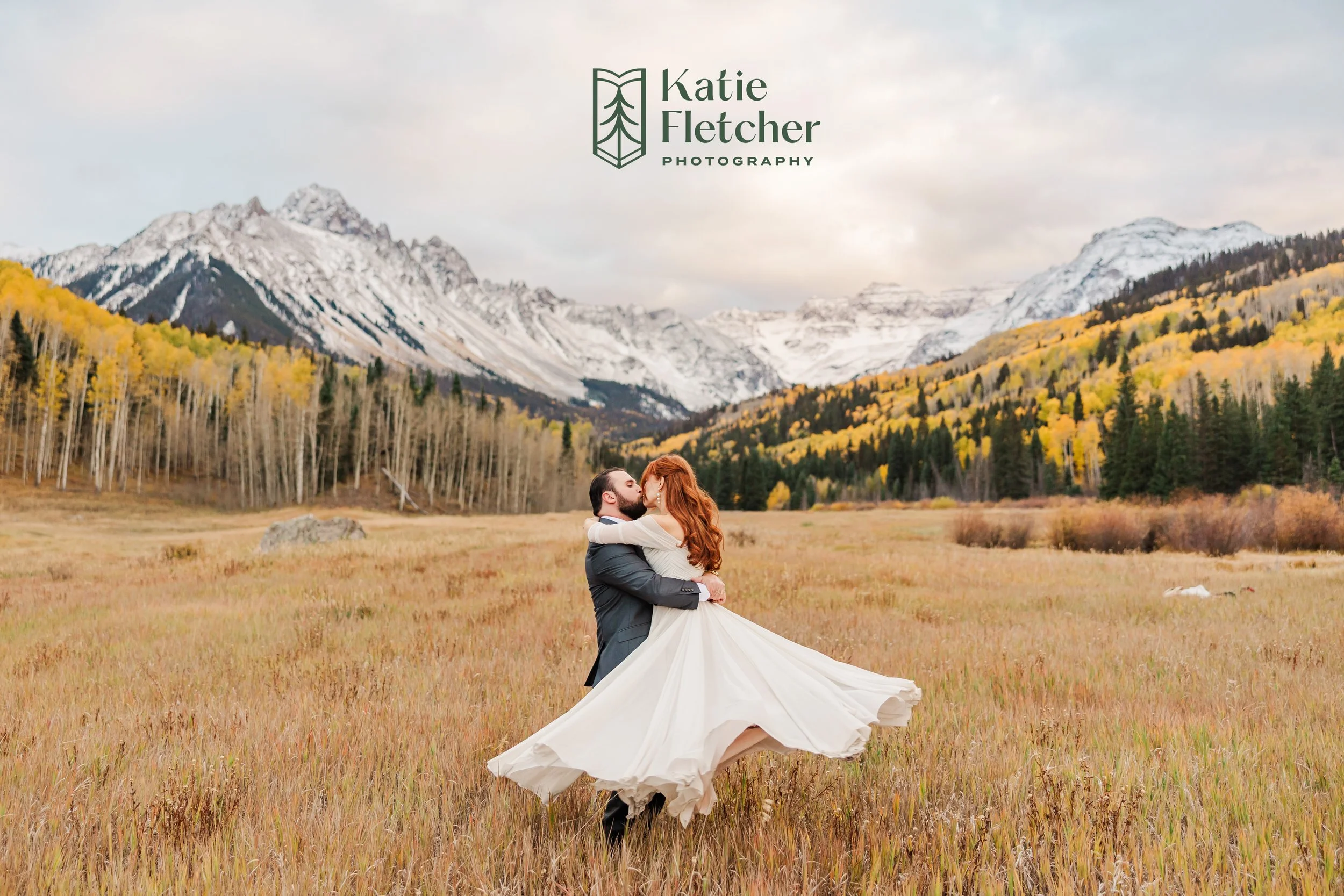 A couple, the man in a black suit and the woman in a white dress, sharing a kiss in a grassy field with mountains, trees, and snow-capped peaks in the background during fall.
