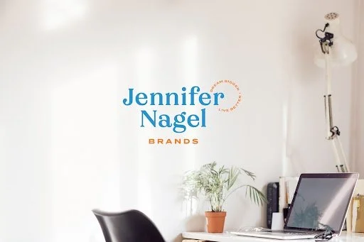 Minimalist office workspace with a laptop, a potted plant, and a black chair in front of a white wall with the logo 'Jennifer Nagel Brands' in blue and orange.