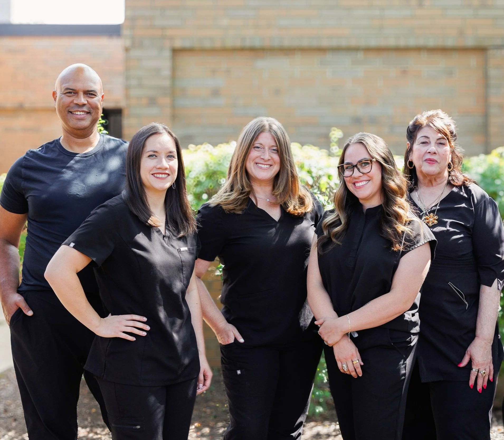 Group of five adults, four women and one man, standing outdoors in front of a brick building and green bushes, smiling at the camera.