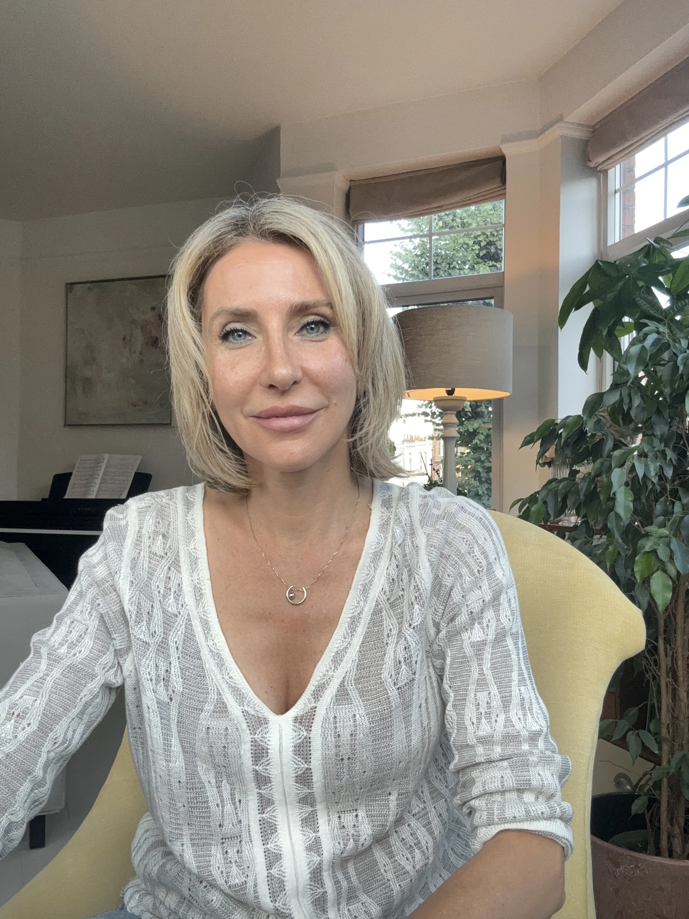 A woman with blonde, shoulder-length hair smiling and sitting in a room with large windows, a tall lamp, and a potted plant, wearing a white patterned top and gold necklace.