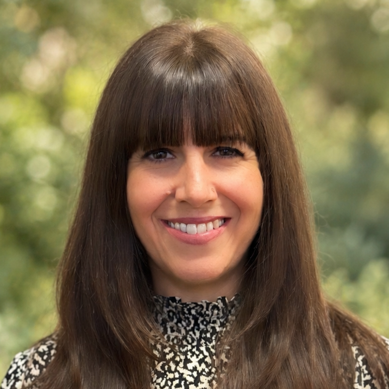Close-up portrait of a woman with straight brown hair and bangs, smiling outdoors with a blurred green natural background.