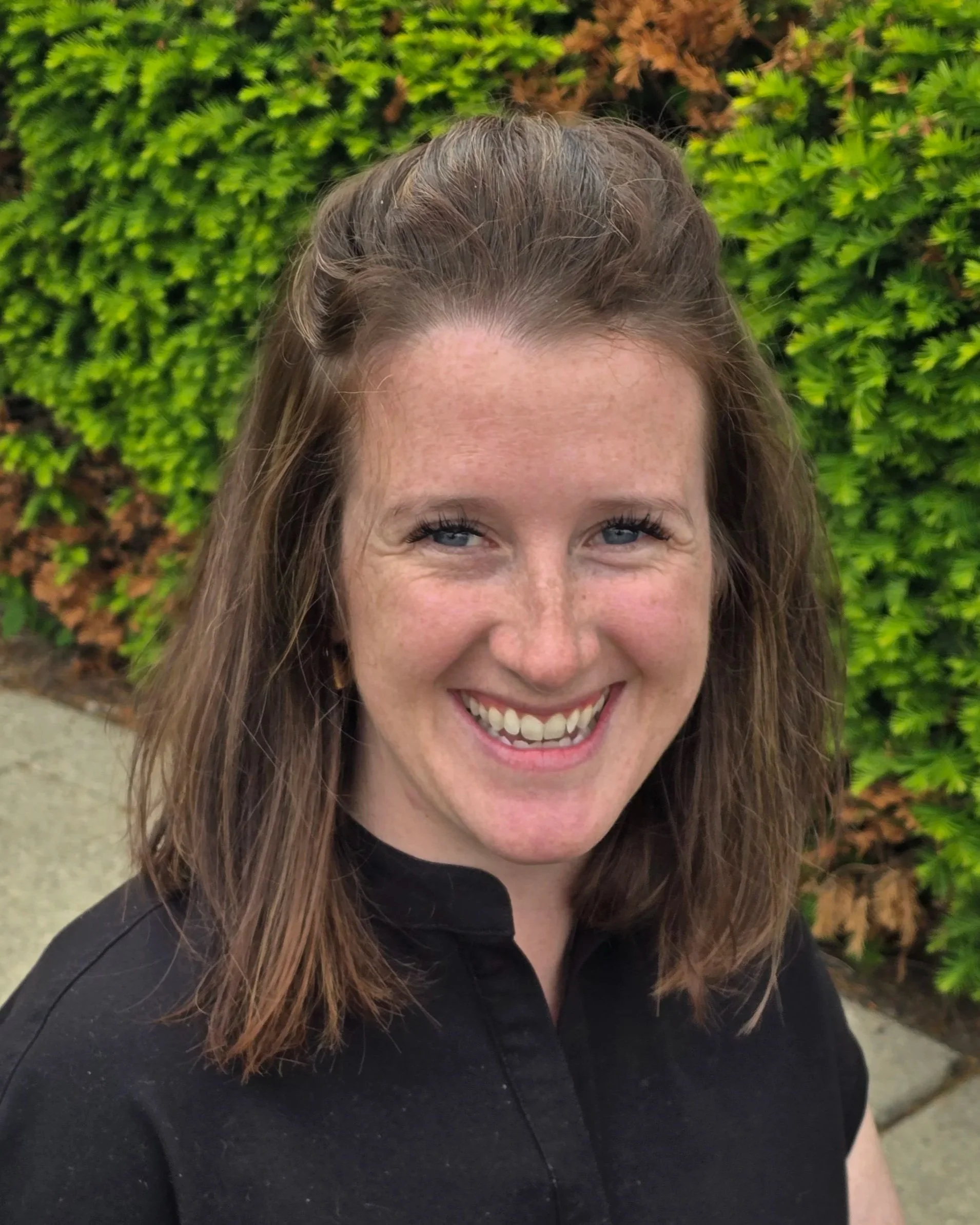 Close-up of a woman with light brown hair, blue eyes, and a big smile, wearing a black top, outdoors with green shrubbery in the background.