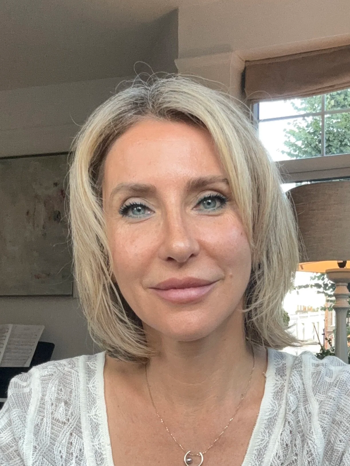 A woman with short blonde hair, blue eyes, wearing a white lace top, and a delicate necklace, sitting inside near a window with a view of trees and buildings outside.