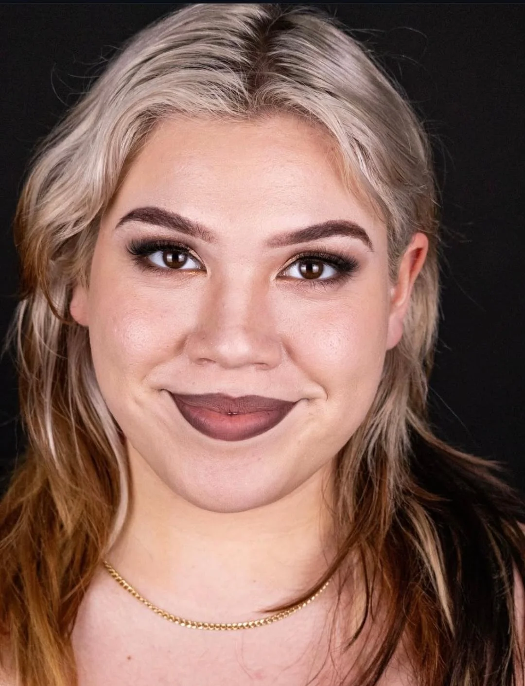 Close-up portrait of a woman with light skin, blonde hair with darker roots, dark brown eyes, and dark lipstick, wearing a gold necklace and makeup, against a black background.
