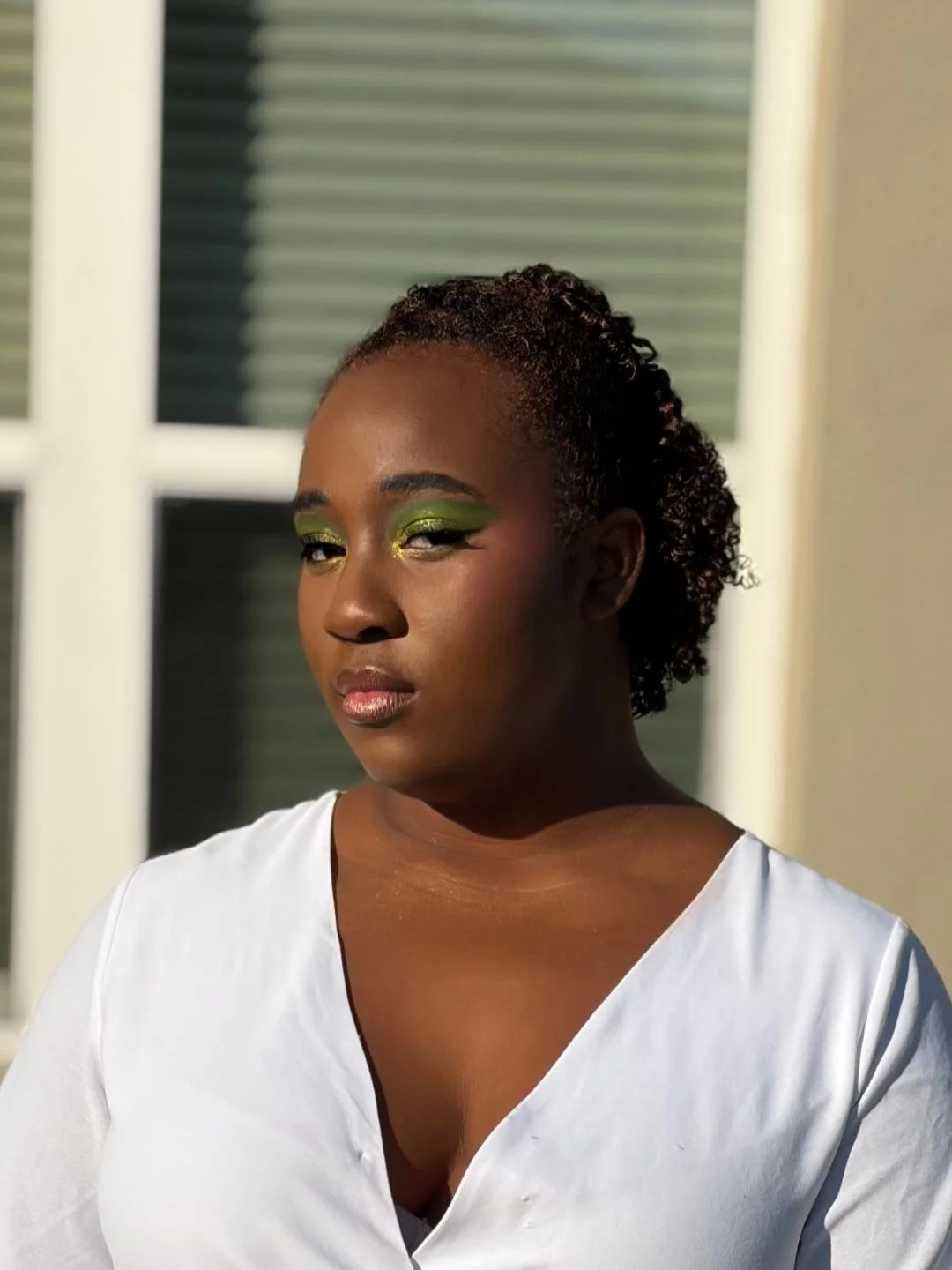 A woman with dark skin and short curly hair, wearing green eye makeup and a white shirt, standing outside near a building with windows.
