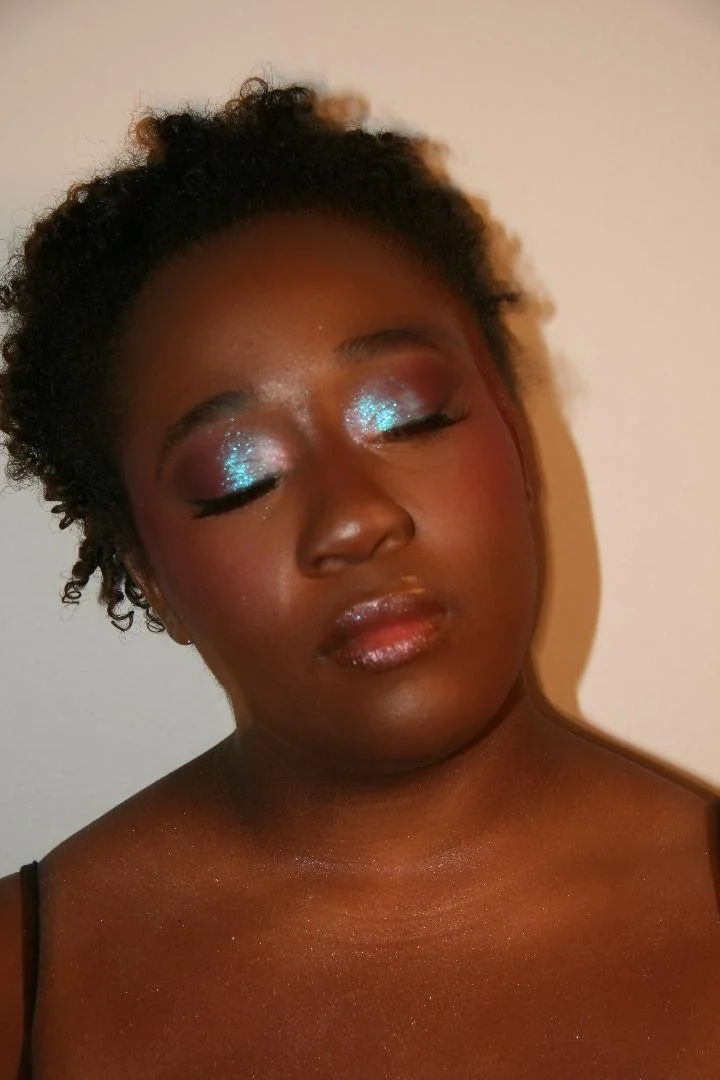 Close-up of a woman with her eyes closed, wearing glittery blue eyeshadow and glossy lips, with natural curly hair and a dark skin tone.