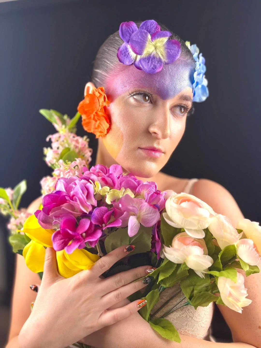 A woman with pastel-colored flowers on her face and head, holding a large bouquet of pink, yellow, white, and purple flowers against a dark background.