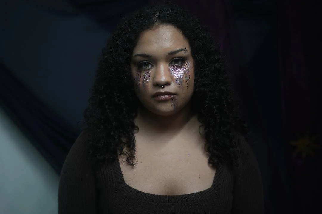 Portrait of a woman with curly black hair and glitter makeup on her face, wearing a black top, against a dark background.