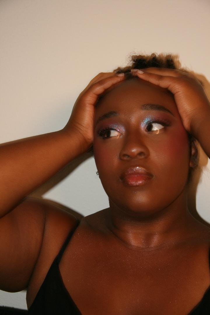 A woman with dark skin, wearing makeup, holding her head with both hands, and looking to the side against a plain wall.