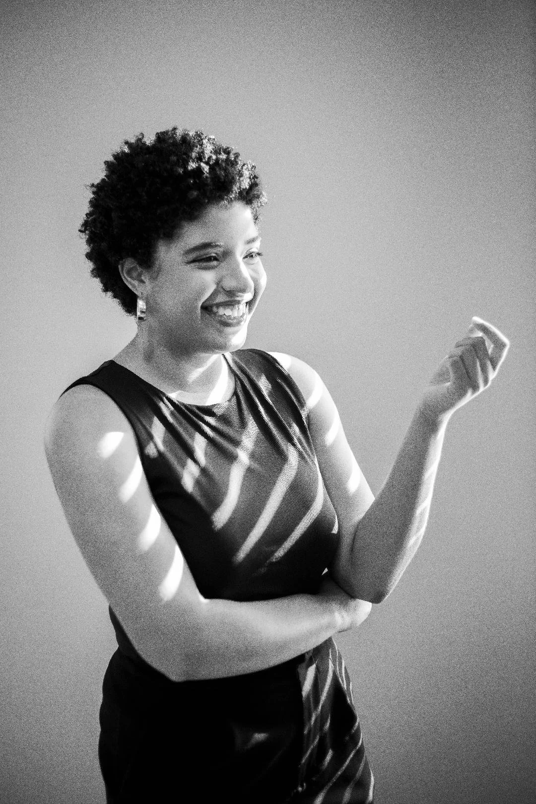 A smiling woman with short curly hair wearing a sleeveless patterned dress, holding a small object in her right hand, with light and shadow lines across her face and body.