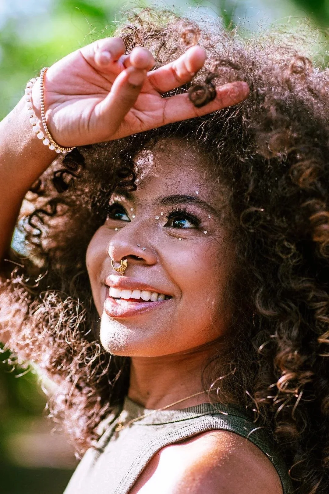 A young woman with curly hair, smiling, with jewelry on her nose, eyebrows, and face, outdoors.