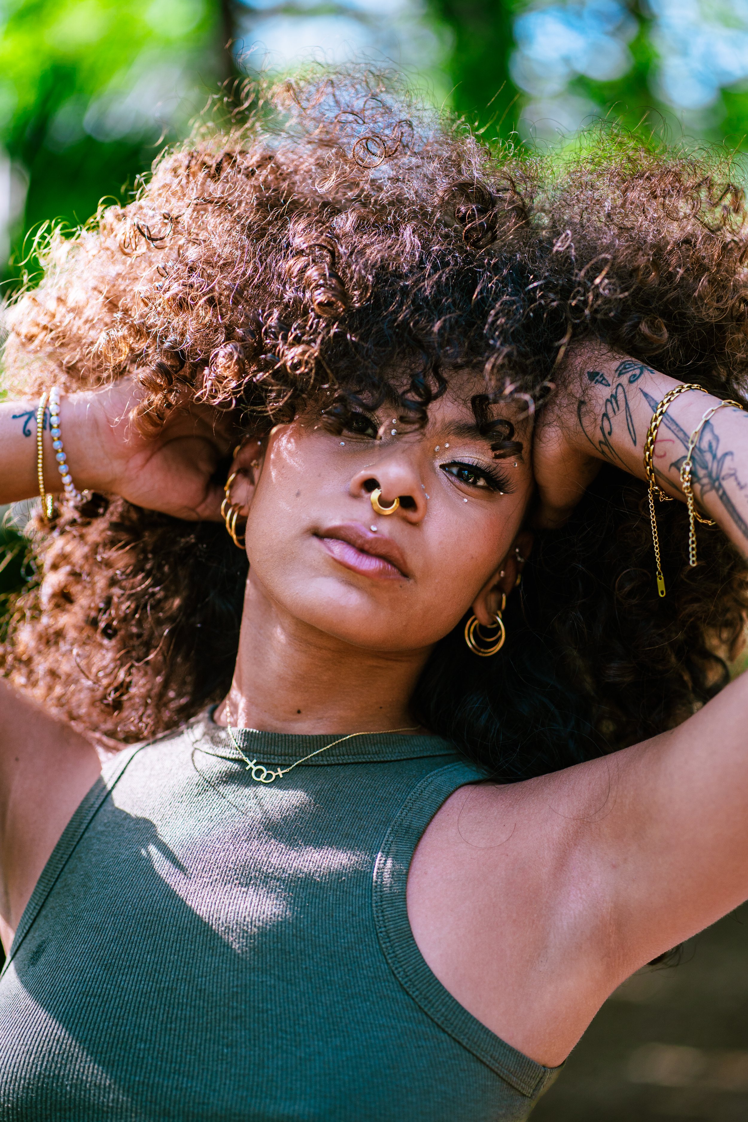 Close-up of a woman with curly hair and tattoos posing outdoors, wearing a green tank top and gold jewelry with a clean edirotial makeup