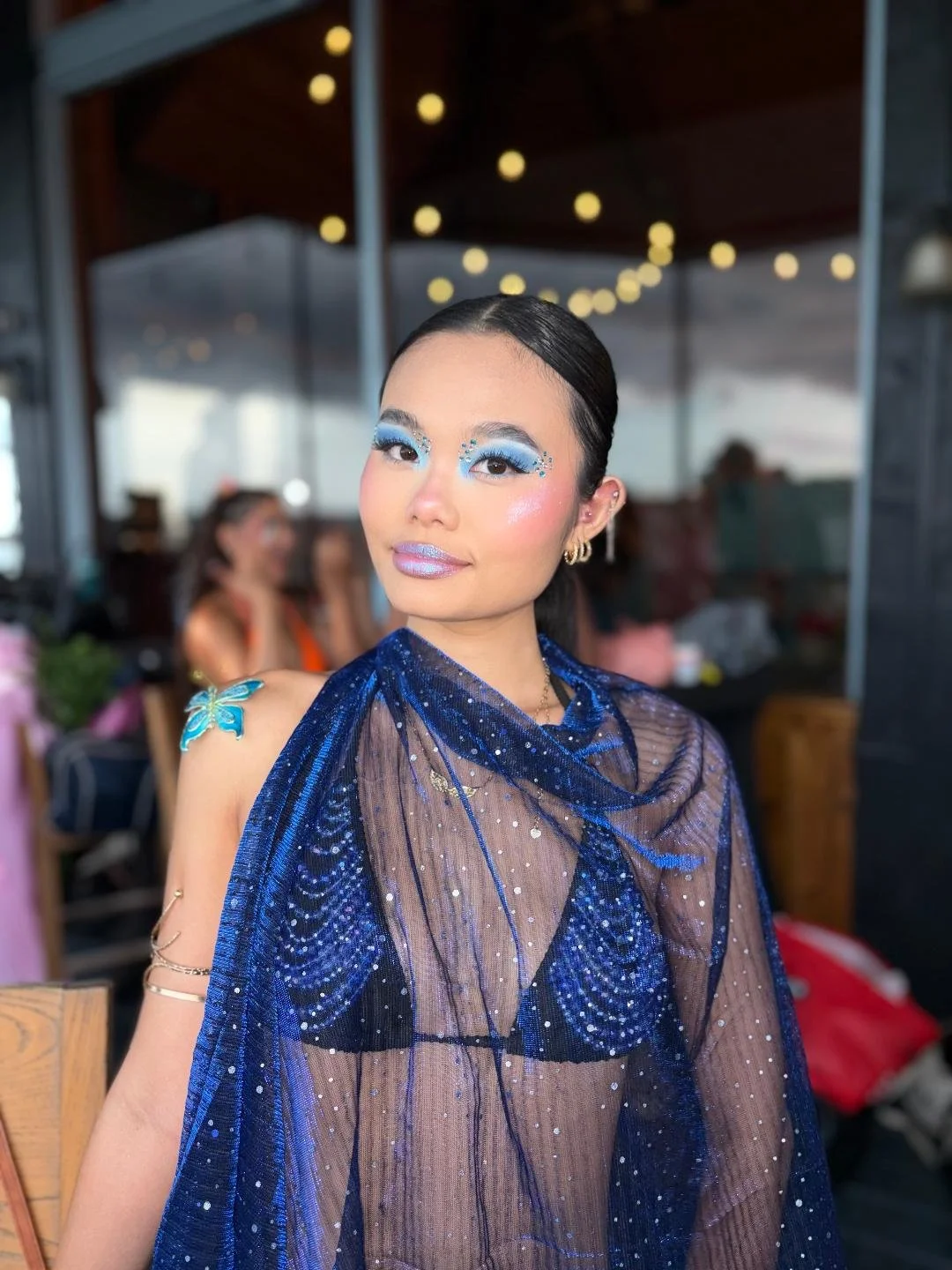 A woman in a sparkly blue shawl and black bikini wearing bright blue makeup and sparkly lips
