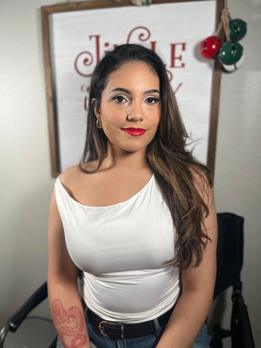 A woman with long brown hair and red lipstick, wearing a white sleeveless top, stands in front of a whiteboard with red and black writing, and a string of red and green Christmas bells hangs on the side.