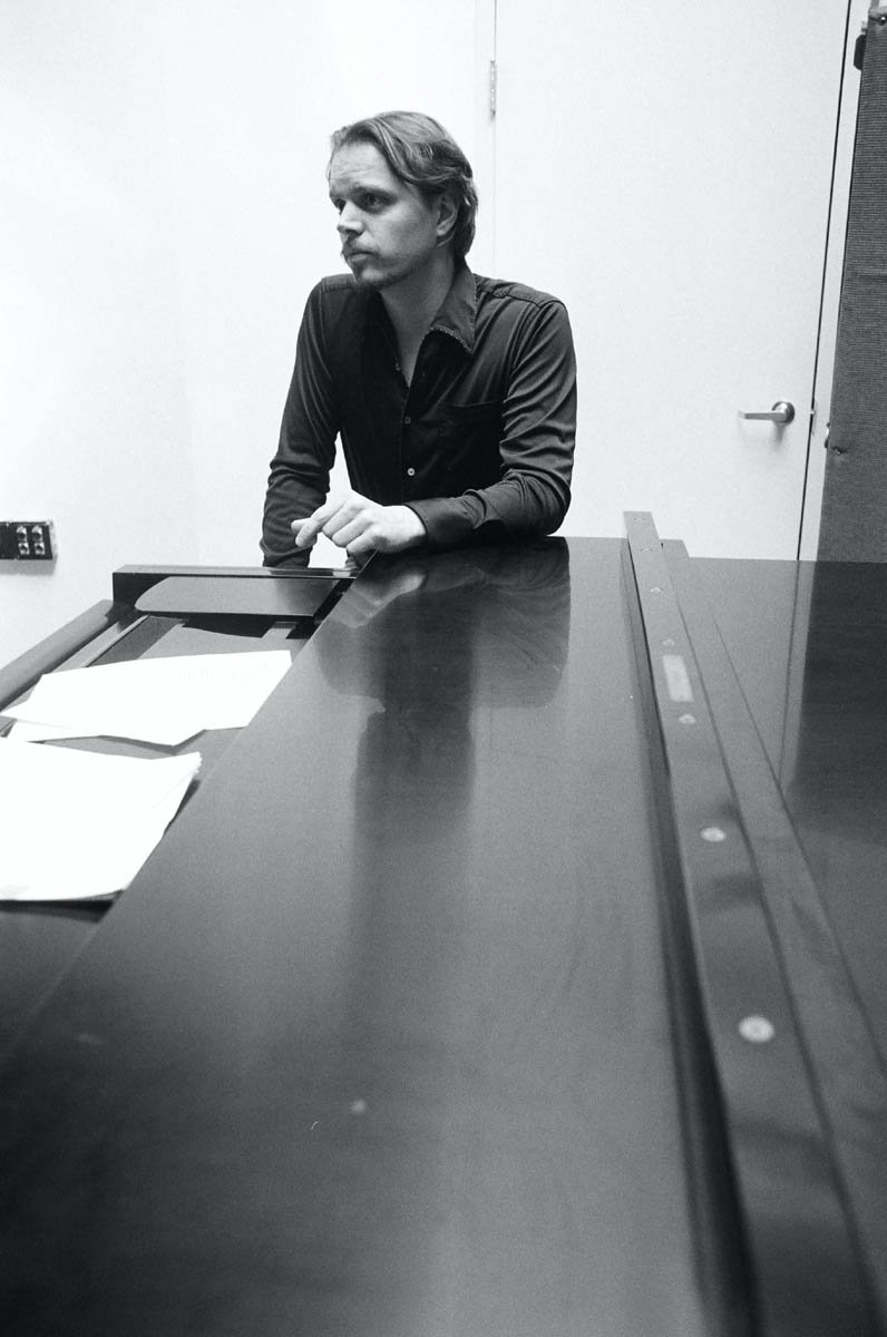 Man with wavy hair and a dark button-up shirt sitting at a desk in a plain room.
