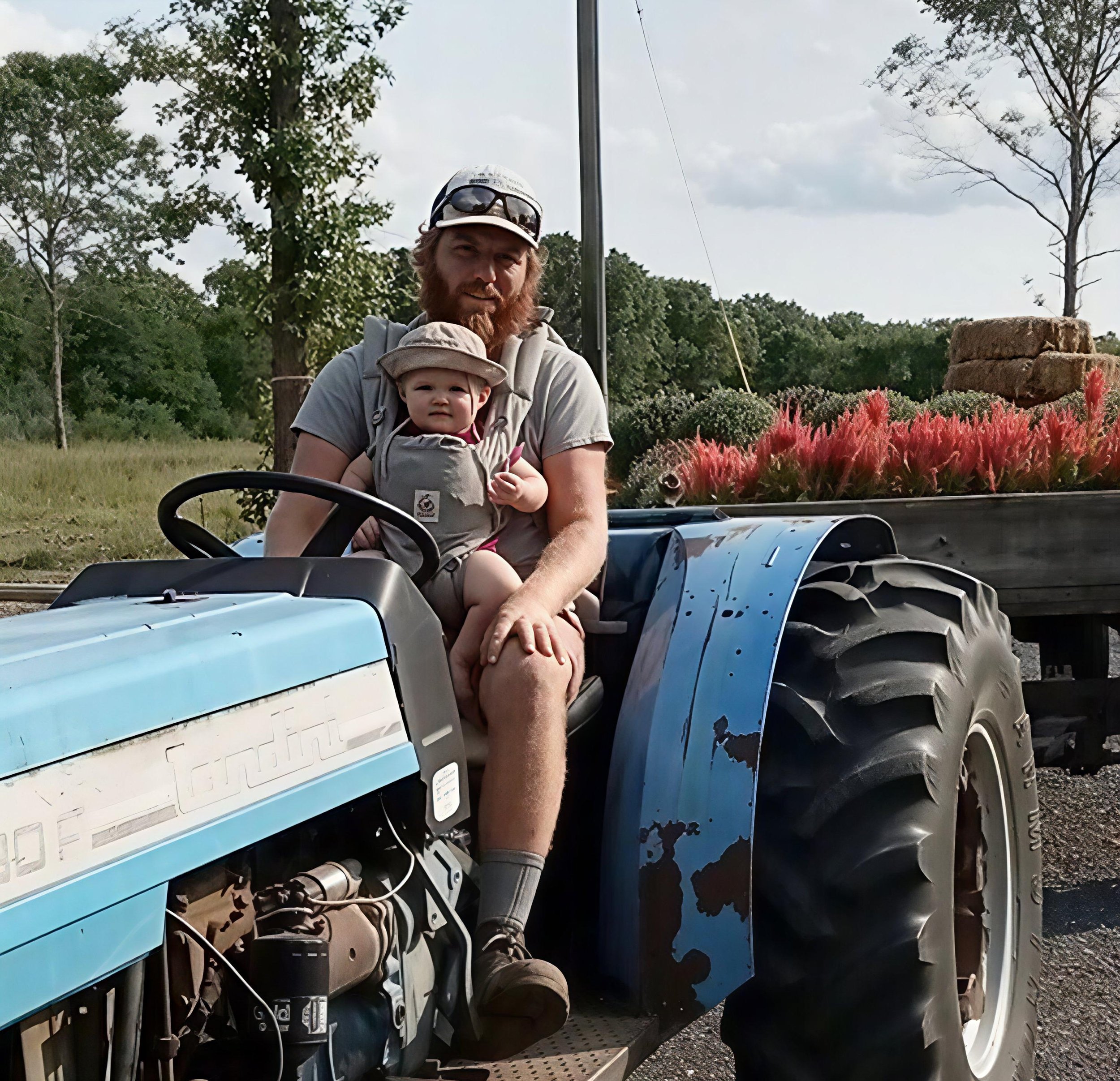A man and a young child sitting on a blue tractor, with the man holding the child while they both face the camera. The child is wearing a hat. The tractor is in an outdoor setting with trees, grass, and a truck bed filled with pink flowers and hay bales in the background.
