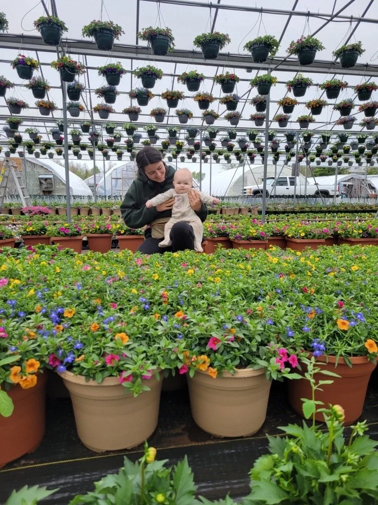 A woman holding a baby in a greenhouse or garden center filled with colorful flowering plants in pots, with hanging plant baskets overhead and a parking lot visible in the background.
